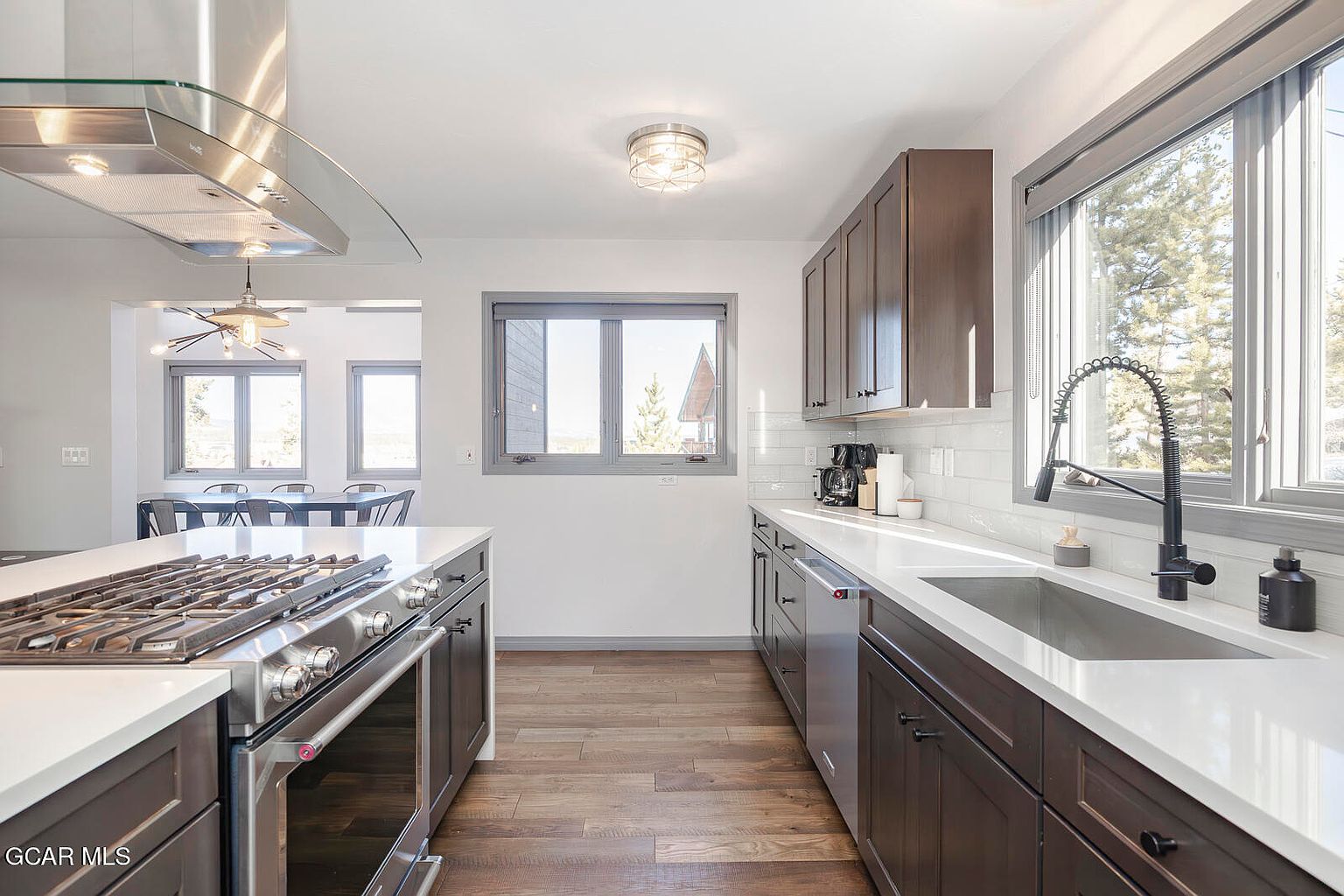 This is a well-lit kitchen featuring dark wood cabinetry, stainless steel appliances, and white countertops. A large island with a gas cooktop is in the foreground, while a sink and window are visible along the back wall. The kitchen has a modern and clean design.