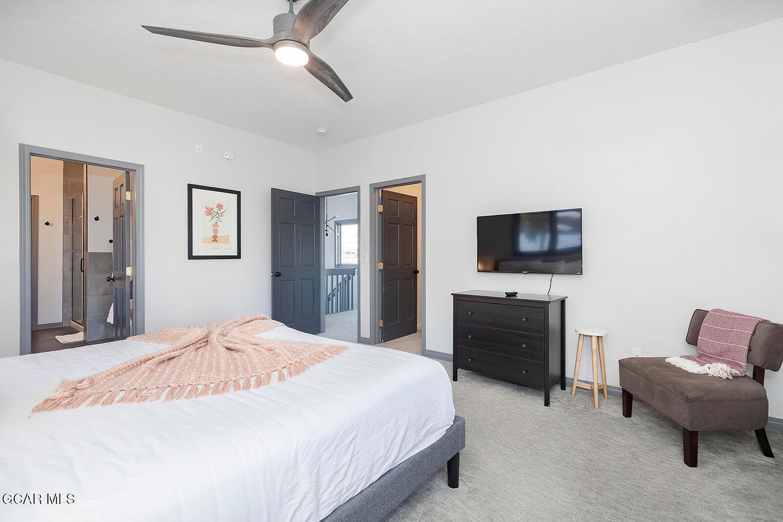 This is a well-lit primary bedroom featuring a bed with a white comforter and a pink throw blanket. The room includes a dark wood dresser with a wall-mounted TV above it, a small stool, and a brown upholstered chair. The walls are painted a neutral color, and there are two doorways leading to other areas of the house.