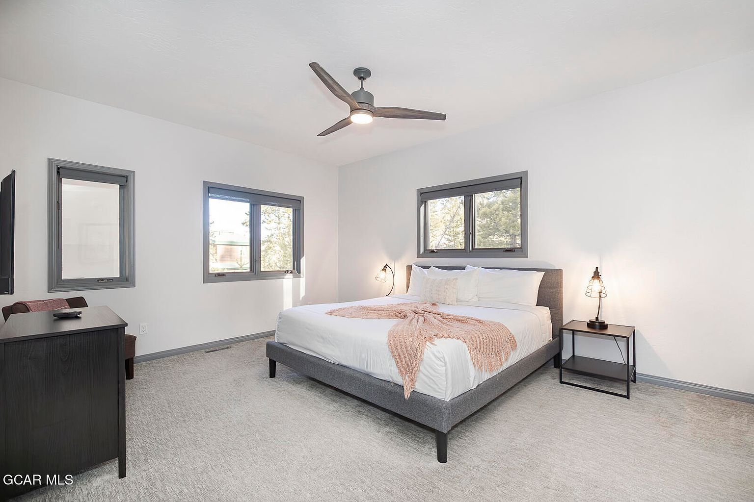 This is a bright and airy primary bedroom featuring a gray platform bed with white linens and a peach-colored throw. The room has three windows with gray trim, allowing natural light to fill the space. A dark wood dresser and a black nightstand with a modern lamp add functional and stylish touches to the room, which is carpeted in a neutral tone.