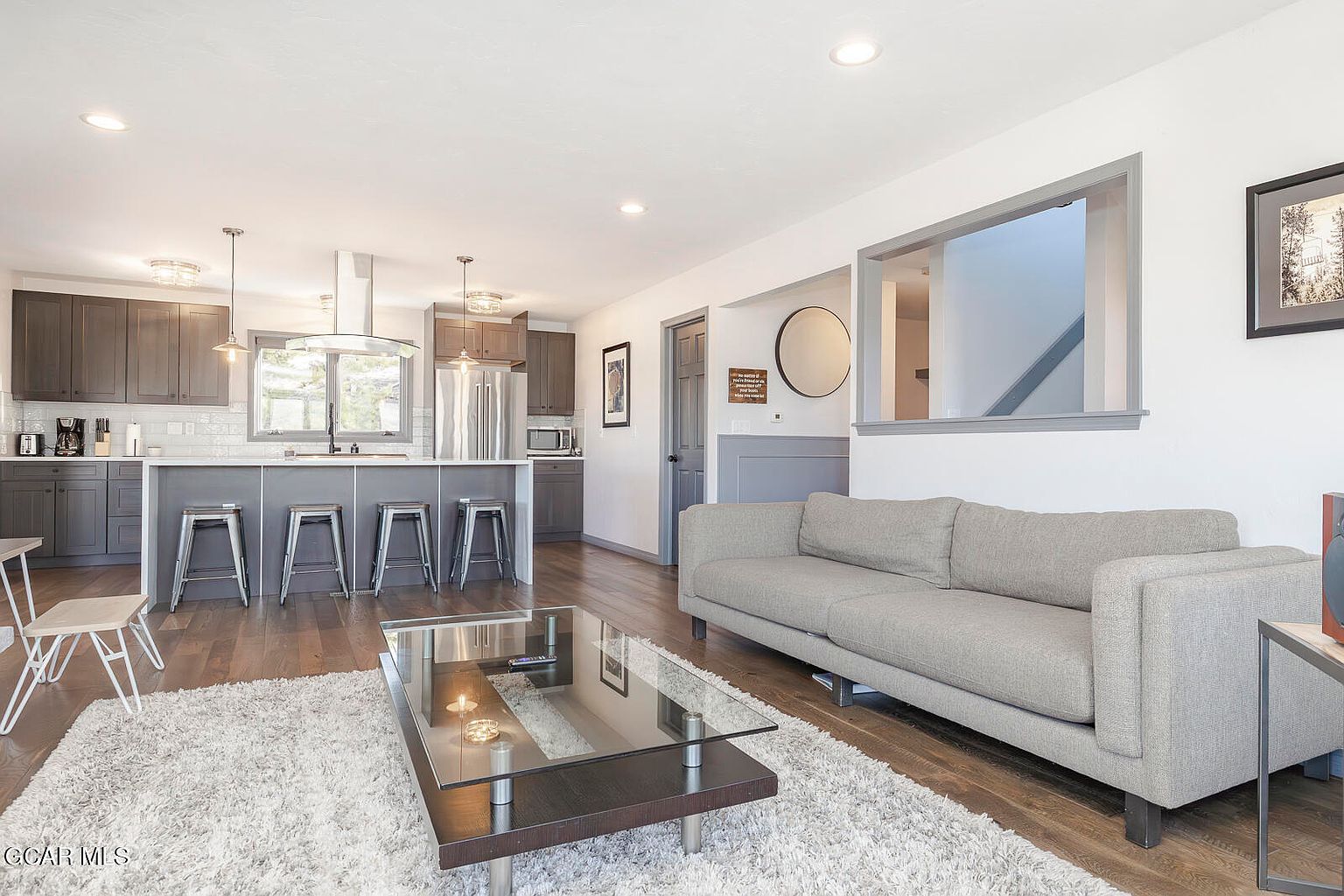 This is an interior shot of a living room and kitchen area. The living room features a gray sofa, a glass-topped coffee table on a white shag rug, and a view into the kitchen. The kitchen has gray cabinets, stainless steel appliances, and a breakfast bar with stools, creating an open and inviting space.