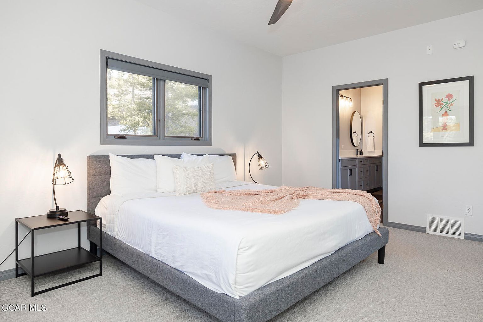 This is a well-lit primary bedroom featuring a gray upholstered bed with white linens and a light pink throw. A black nightstand with a lamp sits beside the bed, and a window provides natural light. An ensuite bathroom is visible through an open doorway, and artwork hangs on the wall, creating a serene and inviting atmosphere.