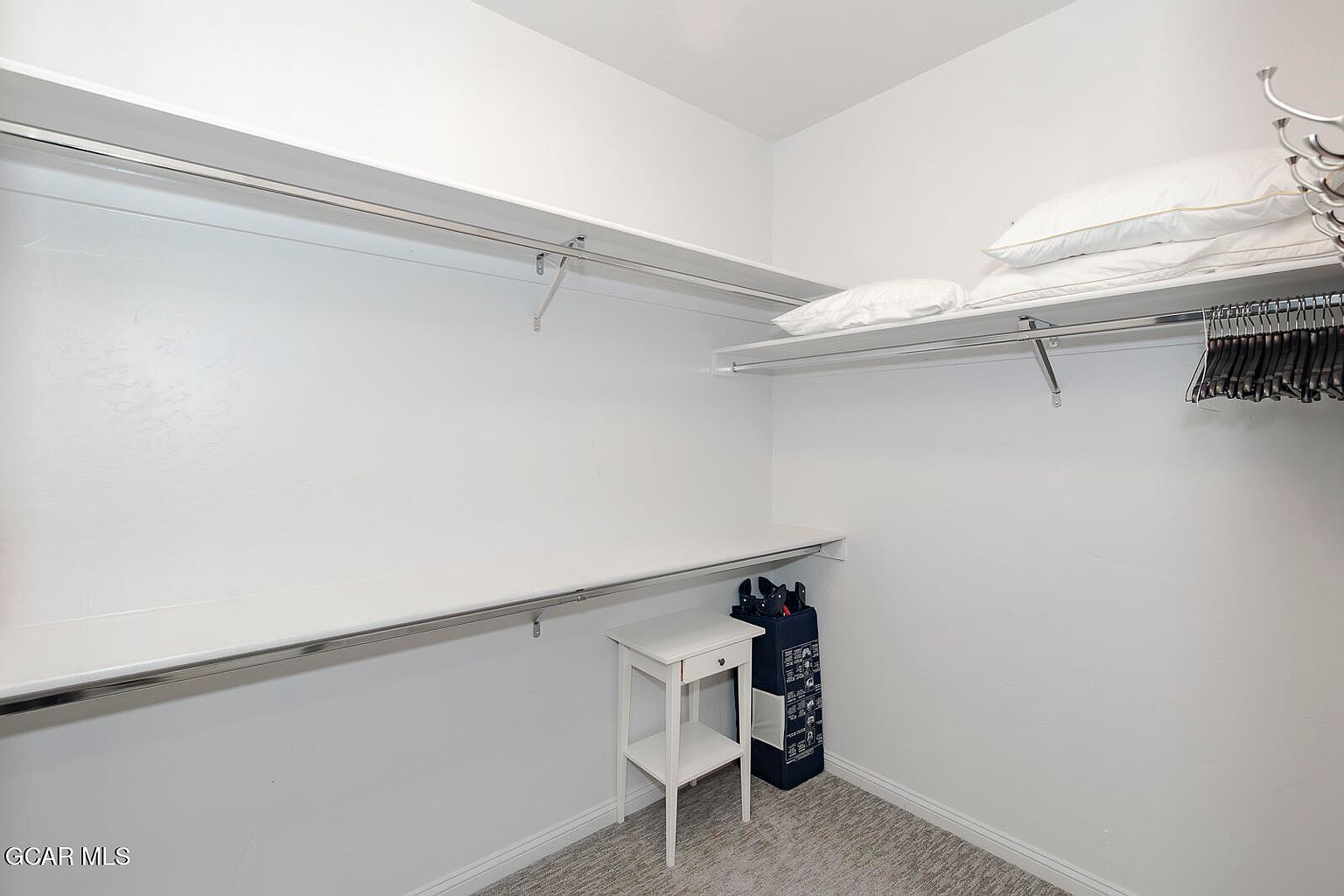 This is a well-organized walk-in closet with white walls, shelving, and a hanging rod. The closet features multiple levels of shelving for storage and a small side table. The overall impression is clean and functional, providing ample space for clothing and accessories.