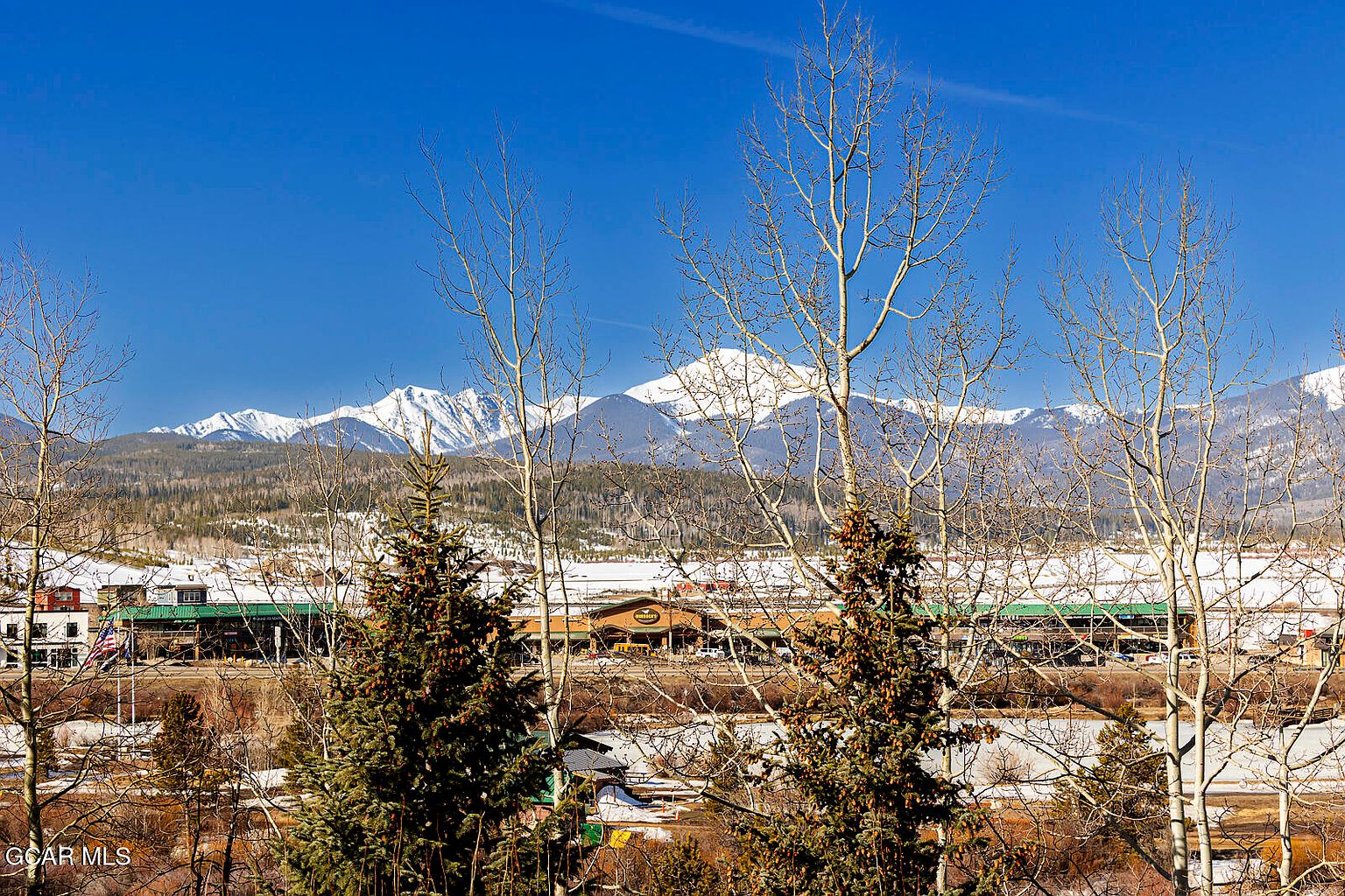 The image showcases a scenic view of a building complex with a green roof, framed by bare trees and evergreen trees in the foreground. Snow-capped mountains rise in the background under a clear blue sky, creating a picturesque and serene setting. The perspective is from an elevated position, offering a wide view of the landscape and the building's relationship to its environment.