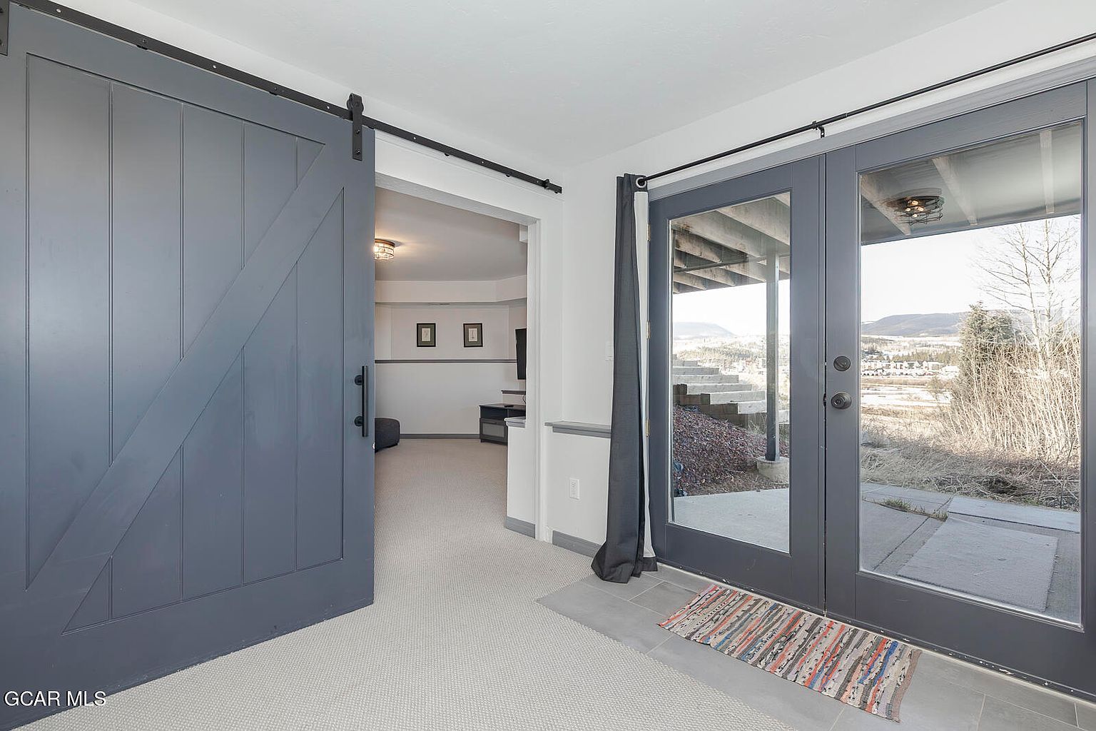The image showcases a basement area with a sliding barn door partially open, leading into another room. A set of glass doors provides access to the exterior, revealing a view of the landscape. The flooring is carpeted, and a small rug lies near the glass doors, creating a cozy and inviting atmosphere.