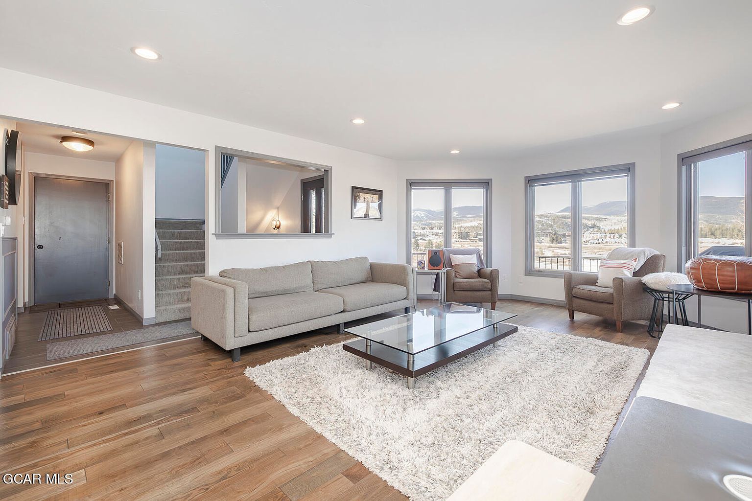 This is an interior shot of a living room featuring a neutral-toned sofa, two armchairs, and a glass-topped coffee table on a plush rug. Large windows offer views of the landscape, and the room is well-lit with recessed lighting, creating a bright and inviting atmosphere. The perspective is from a medium angle, capturing the entire living space.