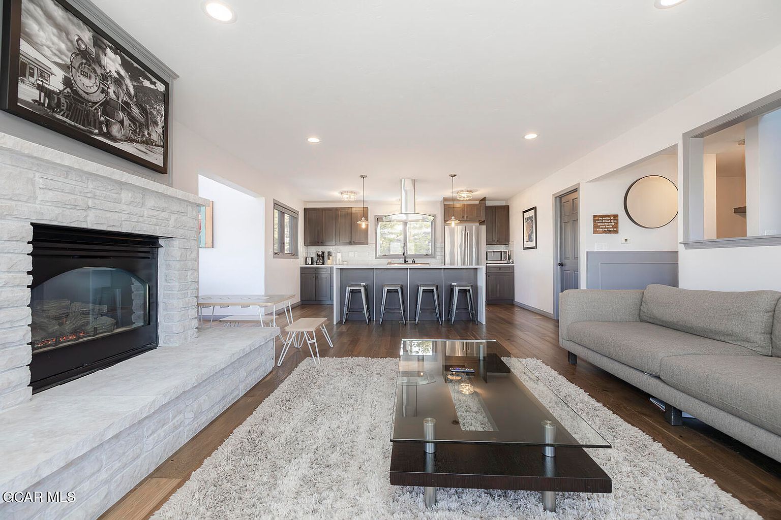 This is an interior shot of a living room that seamlessly flows into a modern kitchen. The living room features a stone fireplace, a plush rug, and a contemporary glass coffee table, while the kitchen boasts dark wood cabinetry, stainless steel appliances, and a central island with bar seating. The open-concept design creates a spacious and inviting atmosphere, perfect for entertaining.