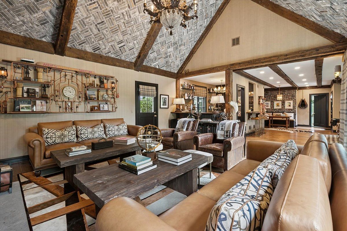 This expansive living room features a vaulted ceiling with exposed wooden beams and a unique patterned tile finish. The space is furnished with two leather sofas and matching armchairs, centered around two rustic wooden coffee tables. A large, intricate copper pipe wall installation serves as a focal point, while an open-concept layout leads into a secondary lounge area with a bar.