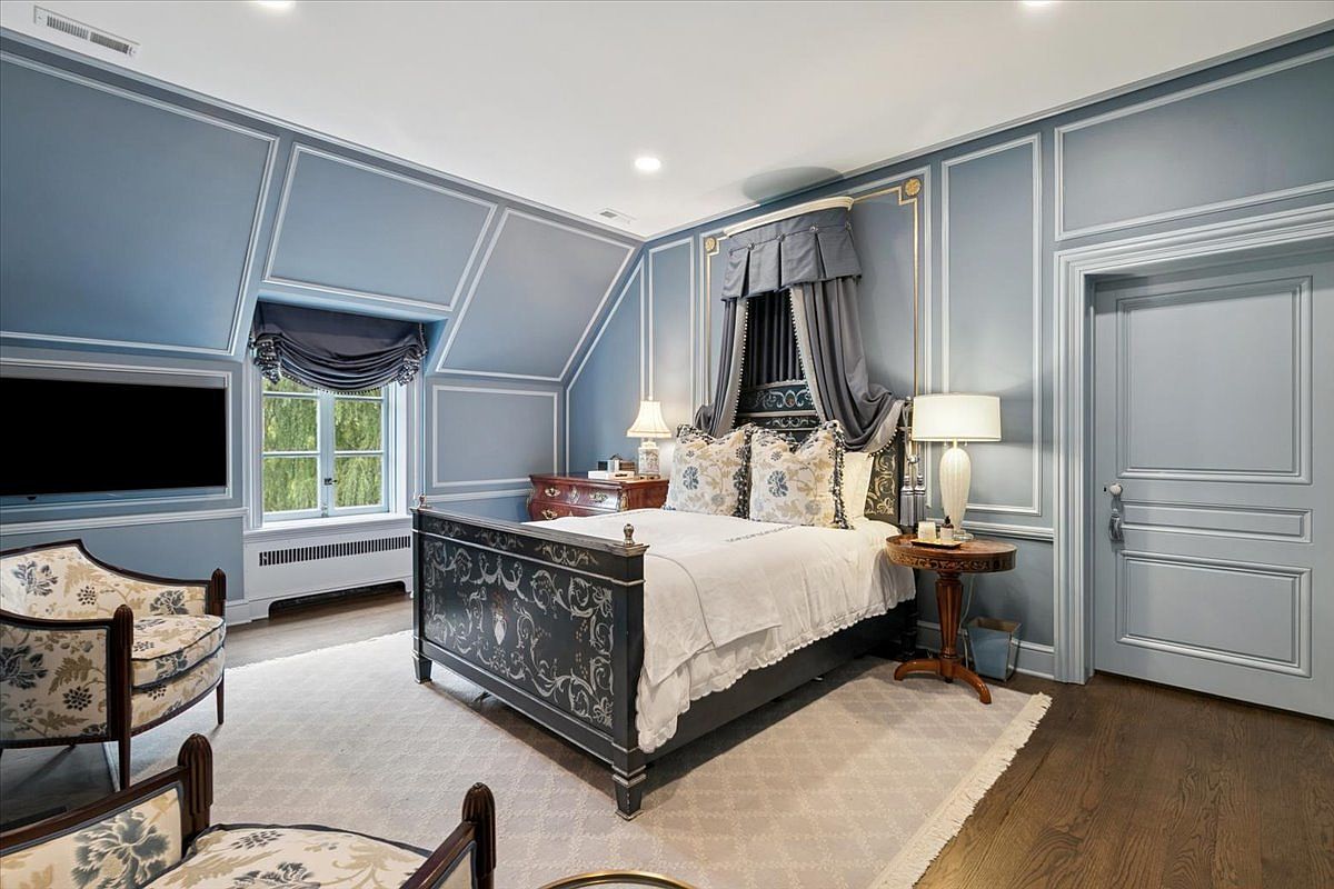 This elegant primary bedroom features sophisticated blue wall paneling and a dramatic canopy bed frame with intricate detailing. The room is furnished with patterned armchairs, a wooden side table with a lamp, and a dresser, all set against a neutral area rug on dark wood flooring. A window with a valance provides natural light, while the overall aesthetic conveys a refined and classic atmosphere.