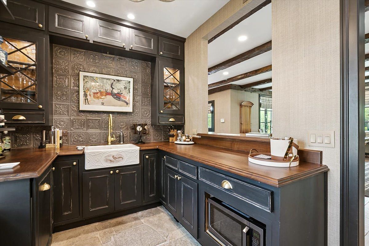 This elegant wet bar or butler's pantry features dark, distressed cabinetry paired with rich, dark wood countertops and a farmhouse-style sink. The backsplash is adorned with decorative metallic-finish tiles, and the space includes a built-in microwave and glass-front upper cabinets. A large opening in the wall provides a view into an adjacent room with exposed ceiling beams, creating an open and sophisticated atmosphere.