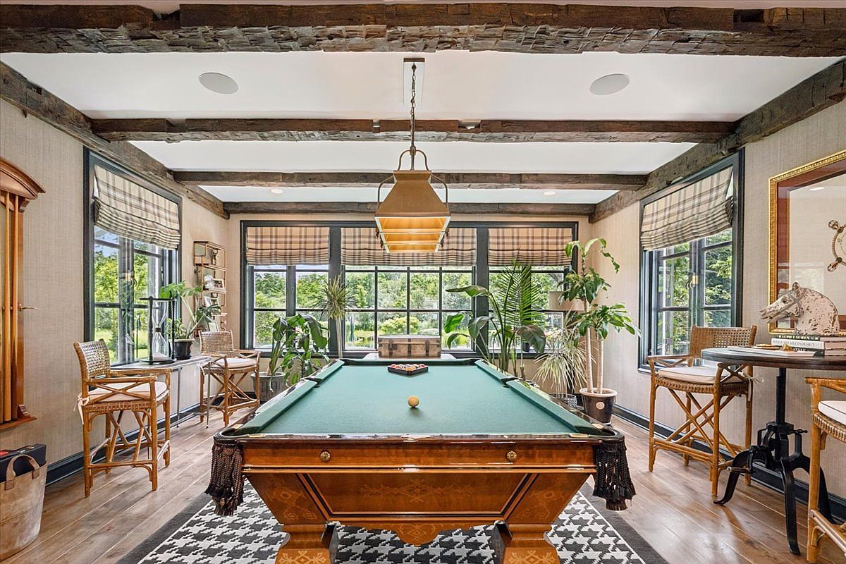 This sophisticated game room features a central billiard table as the focal point, set against a backdrop of large windows with plaid roman shades. The space is defined by rustic exposed wooden ceiling beams and a houndstooth-patterned area rug, creating a refined, classic aesthetic. Wicker-style bar stools and lush potted plants add texture and a touch of warmth to the room's elegant, traditional design.