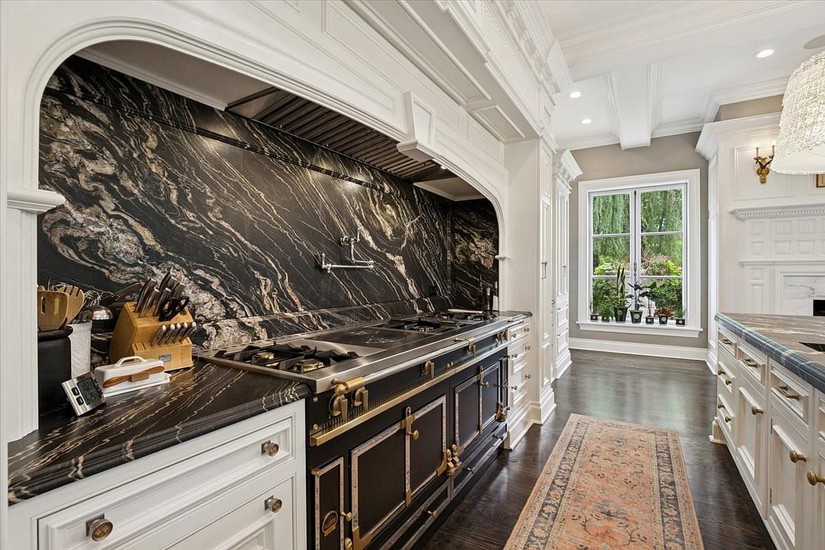 This elegant kitchen features a striking black stone backsplash and countertops with dramatic gold veining, paired with classic white cabinetry and dark wood flooring. A professional-grade black and gold range serves as the centerpiece, set beneath a custom-built range hood. The perspective captures the length of the kitchen, leading the eye toward a window that brings in natural light and views of the greenery outside.