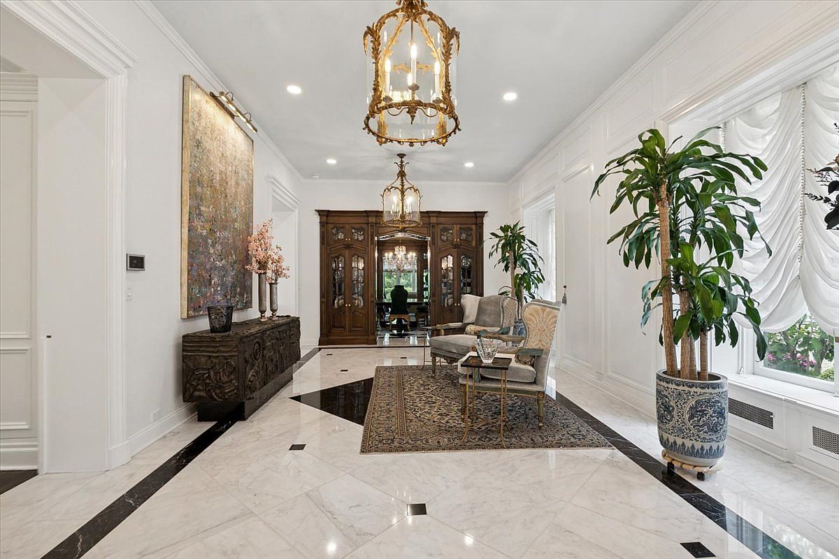 This grand foyer features polished stone flooring with a dark geometric border and a central patterned area rug. The space is illuminated by two ornate gold chandeliers and includes a large abstract painting above a dark, intricately carved console table. Two elegant armchairs are positioned near a large window draped in soft, gathered curtains, while a tall potted plant adds a touch of greenery to the sophisticated, formal entryway.