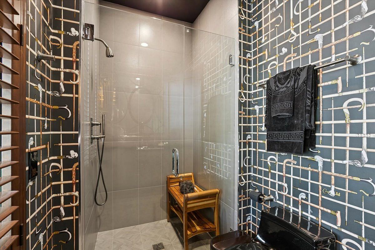 This bathroom features a bold, patterned wall tile with a grid design and umbrella motifs, creating a unique and artistic aesthetic. A glass-enclosed shower area includes a wooden bench, while a dark-toned toilet and a towel rack with branded towels complete the space. The perspective is a straight-on shot that highlights the contrast between the intricate wall patterns and the sleek, modern shower fixtures.