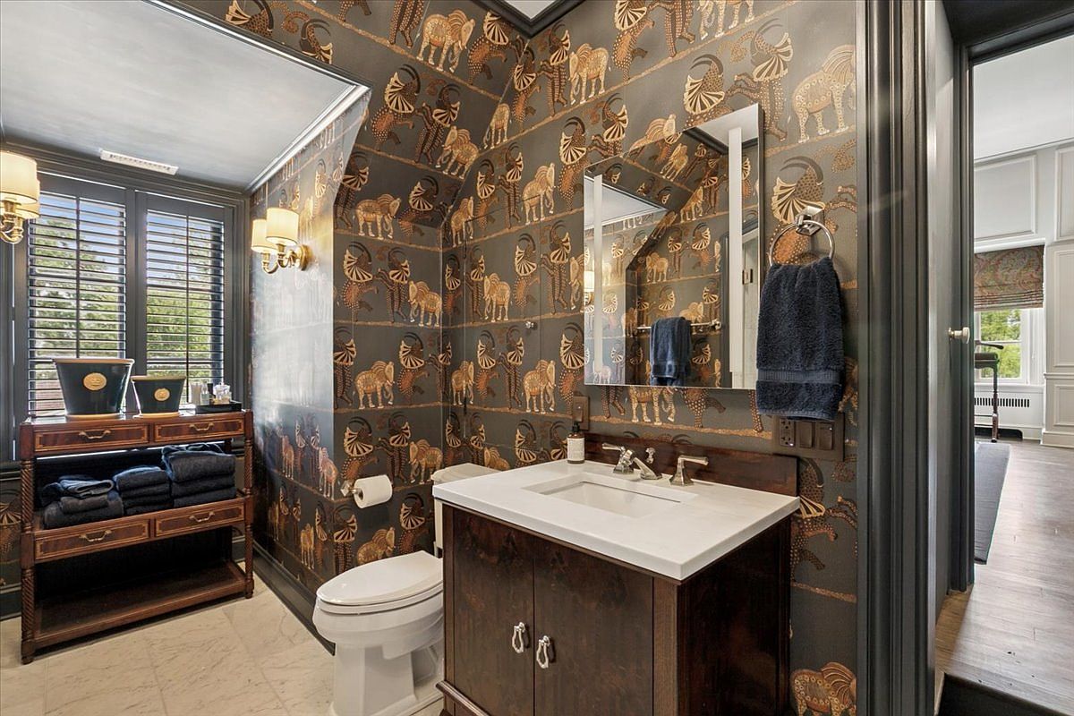 This powder room features bold, animal-patterned wallpaper in dark tones, creating a dramatic and sophisticated aesthetic. The space includes a dark wood vanity with a light-colored stone countertop, a wall-mounted mirror, and a matching open shelving unit for storage. The room is illuminated by wall-mounted sconces and provides a view into an adjacent hallway, emphasizing the cohesive dark trim throughout the home.