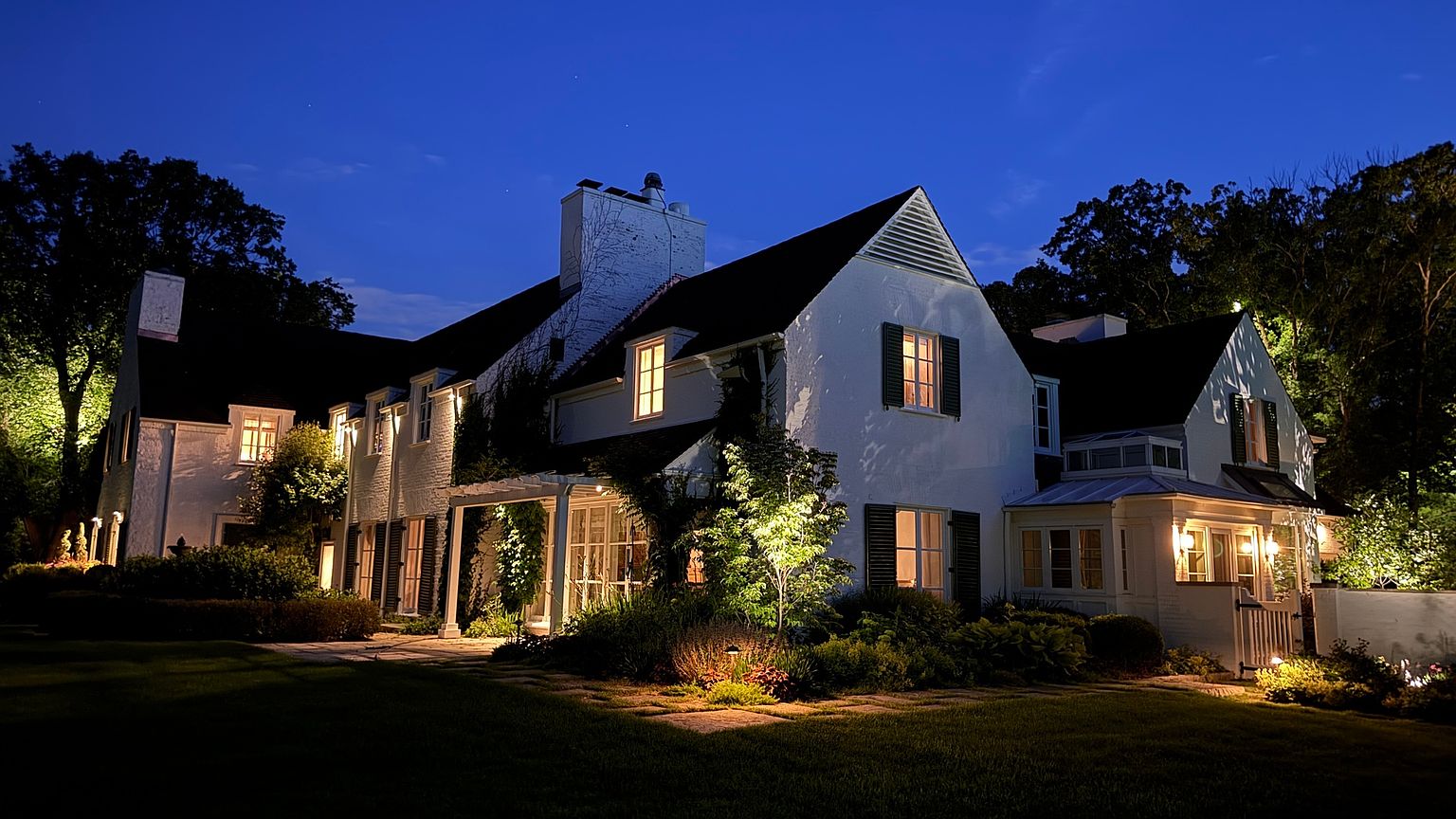 This elegant, white-painted brick home is captured at twilight, showcasing a warm and inviting atmosphere with illuminated windows and landscape lighting. The rear facade features a charming pergola-covered patio area, lush garden beds, and a well-manicured lawn that extends toward the surrounding mature trees. The cinematic perspective highlights the architectural character of the home, emphasizing its classic design and serene evening ambiance.