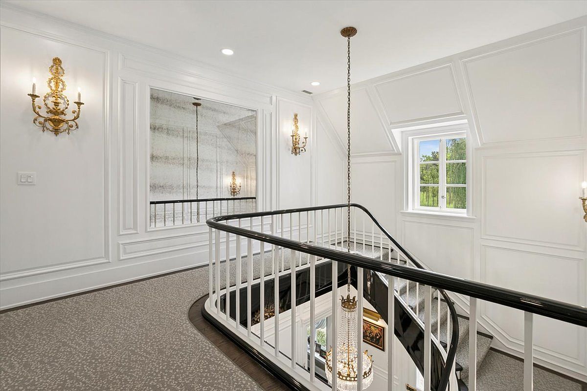 This elegant landing area features a curved staircase with a polished dark handrail and white balusters, leading down to a lower level. The space is adorned with classic wall molding, ornate gold wall sconces, and a large, antique-style mirror that reflects the chandelier hanging above the stairwell. A window provides natural light, while the neutral patterned carpet and crisp white walls create a sophisticated and airy atmosphere.
