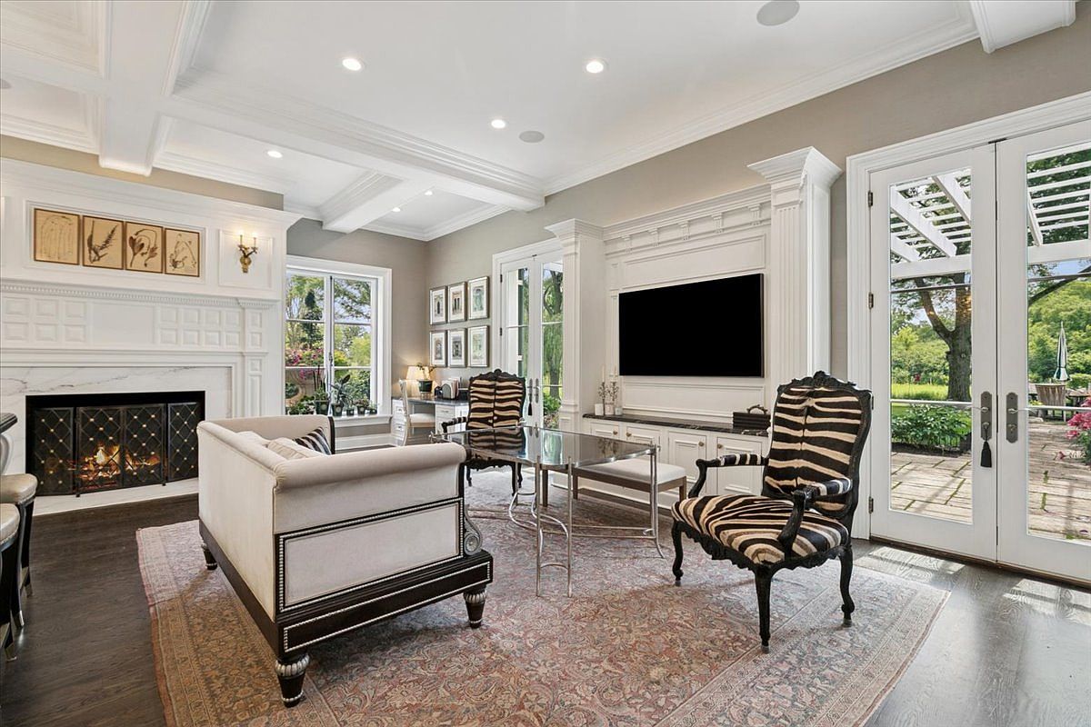 This elegant living room features a sophisticated design with a neutral color palette, dark hardwood floors, and a large patterned area rug. The space is anchored by a fireplace with a decorative mantel and a wall-mounted television above a built-in media console, while glass-paned double doors provide a seamless transition to the outdoor patio. The room is furnished with a plush sofa and two distinctive zebra-print accent chairs, creating a refined and inviting atmosphere.