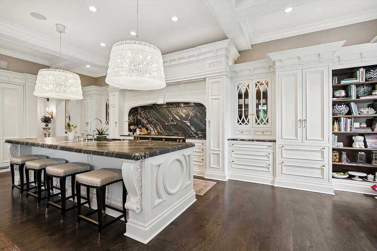 This elegant, high-end kitchen features a large central island with a dark stone countertop and ornate white cabinetry. The space is illuminated by two large, decorative pendant lights and includes a built-in bookshelf and glass-front display cabinets. The dark wood flooring contrasts beautifully with the bright white woodwork, creating a sophisticated and luxurious atmosphere.
