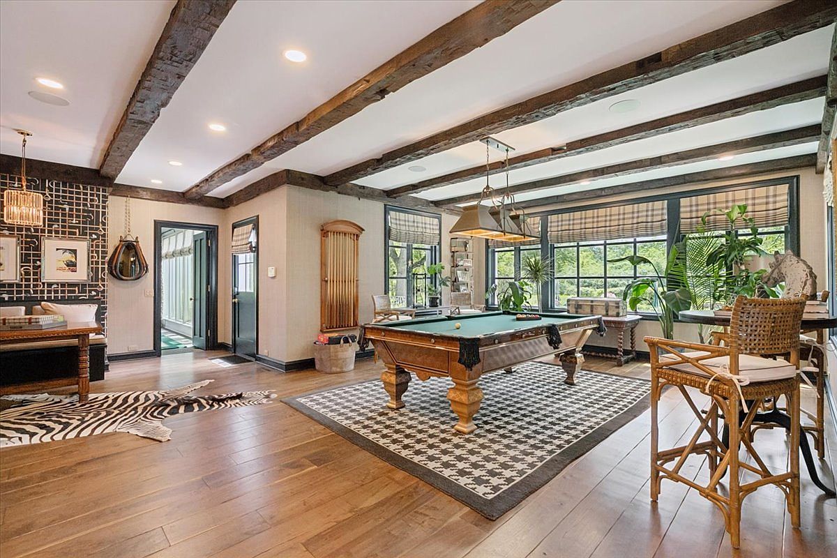 This spacious game room features a prominent pool table centered on a houndstooth-patterned area rug, set against a backdrop of exposed wooden ceiling beams. The room is accented with a zebra-print rug, a unique geometric wall pattern, and large windows that provide ample natural light and views of the outdoors. The space is furnished with a rattan chair and a built-in bench, creating a sophisticated yet relaxed atmosphere for entertainment.