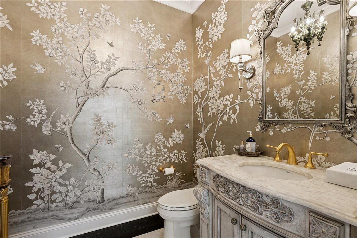This elegant powder room features ornate, chinoiserie-style wallpaper with metallic gold tones and intricate botanical illustrations. The space is anchored by a stone-topped vanity with a carved wooden base and a gold-toned faucet, complemented by a decorative, vintage-inspired mirror and a matching wall sconce. A white toilet is positioned against the patterned wall, creating a sophisticated and luxurious atmosphere.