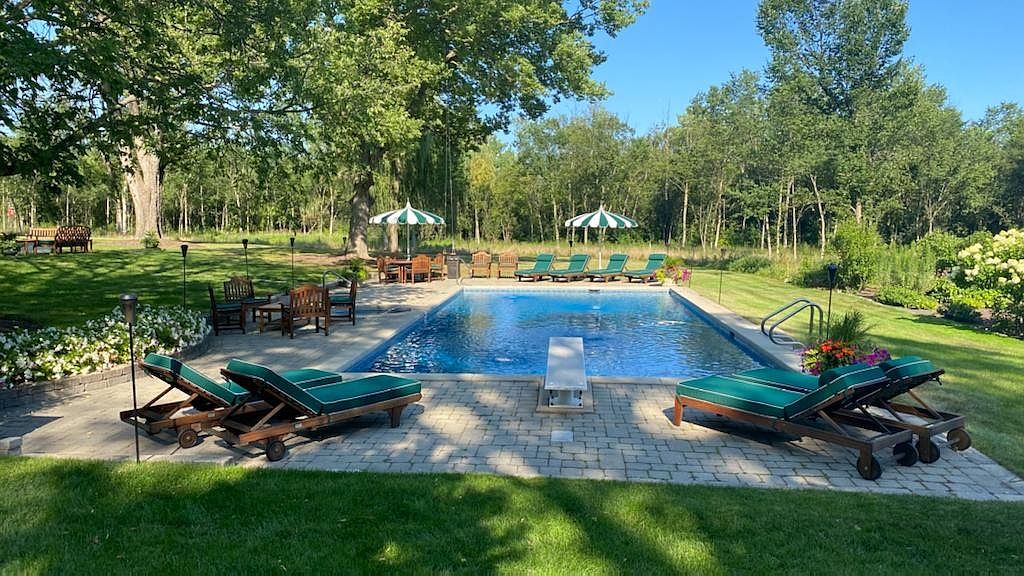 This expansive backyard features a large, rectangular swimming pool as the centerpiece, complete with a diving board and surrounding stone patio. Multiple lounge chairs with green cushions are arranged around the pool, complemented by two striped umbrellas and a dining area set under the shade of mature trees. The property is bordered by lush green lawns and dense woodland, creating a private and serene outdoor retreat.