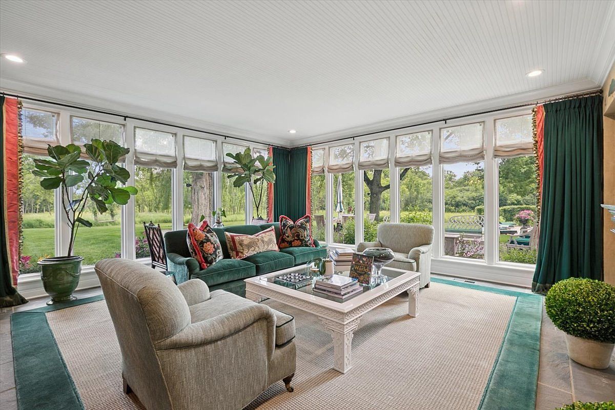 This bright sunroom features a wall of large windows that provide expansive views of the lush green backyard. The space is furnished with a deep green velvet sofa, two upholstered armchairs, and a central white coffee table, all arranged on a textured area rug. The room is accented with potted plants and elegant green curtains, creating a serene and inviting atmosphere.
