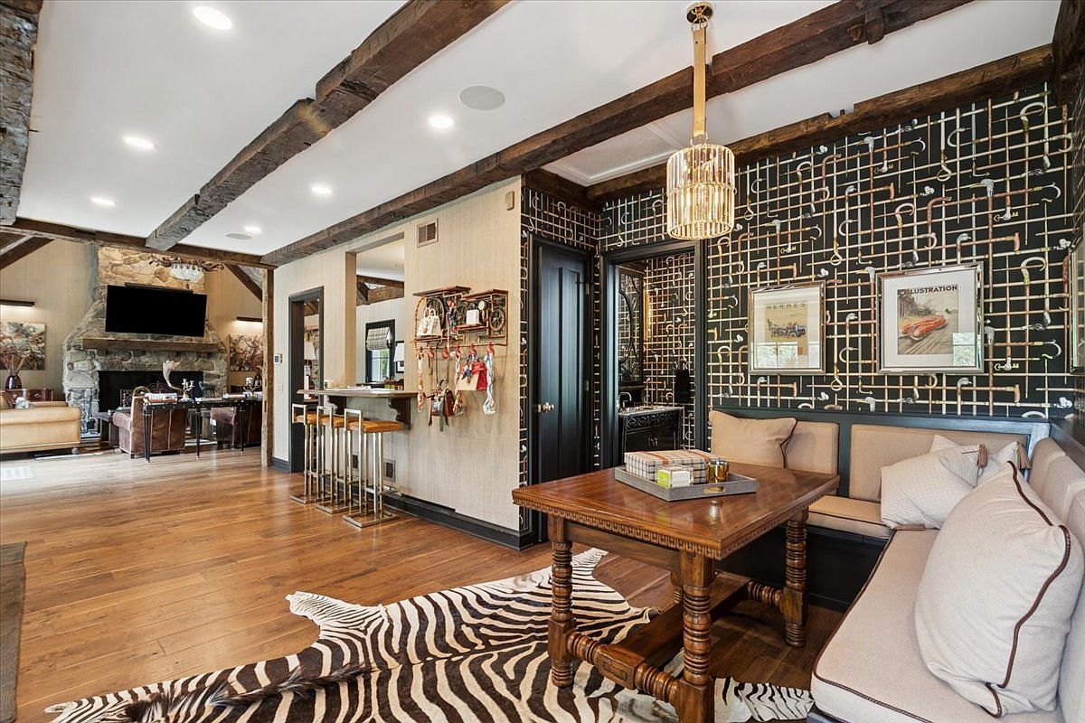 This open-concept dining and living area features a rustic aesthetic with exposed dark wood ceiling beams and warm wood flooring. The dining space includes a wooden table with bench seating, set against a unique wall adorned with a grid of copper-toned pipes and framed artwork. A zebra-print rug anchors the space, while a nearby bar area with stools and a stone-clad chimney breast in the adjacent living room complete the sophisticated, eclectic design.