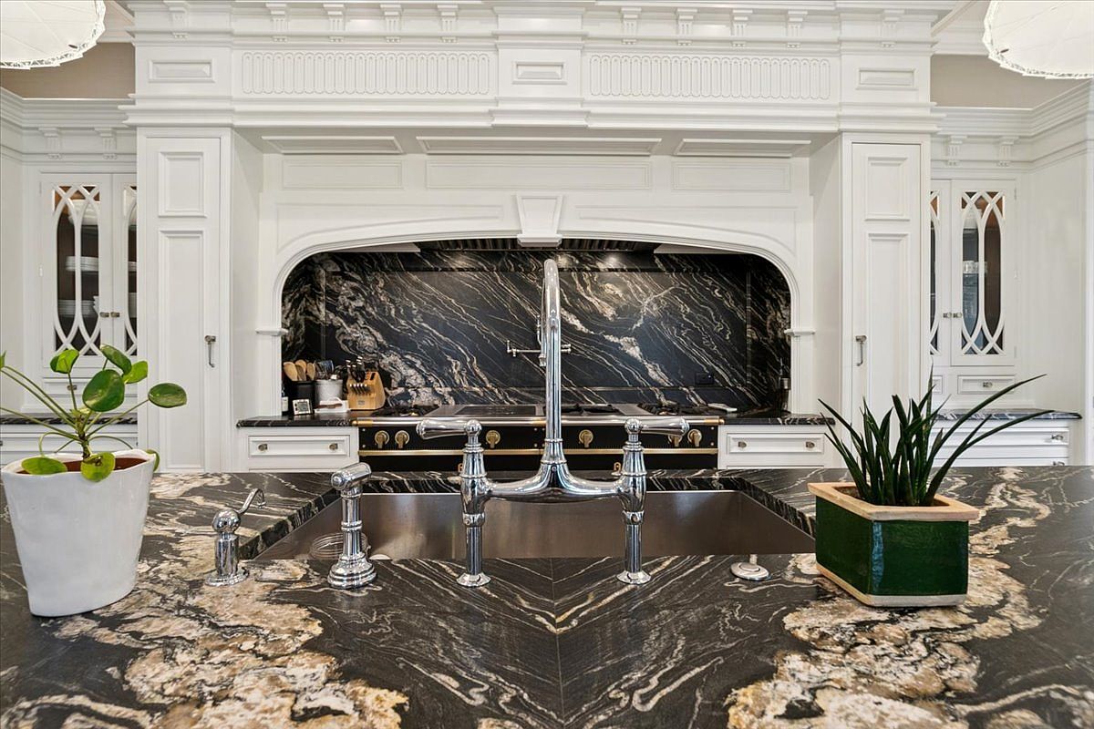 This elegant kitchen features a prominent central island with a striking dark, veined stone countertop and a large stainless steel sink with a polished chrome faucet. Behind the island, a professional-grade range is set into a custom white cabinetry surround, complemented by a matching dark stone backsplash. The perspective is centered on the sink, highlighting the sophisticated contrast between the white millwork and the dramatic, dark stone surfaces.