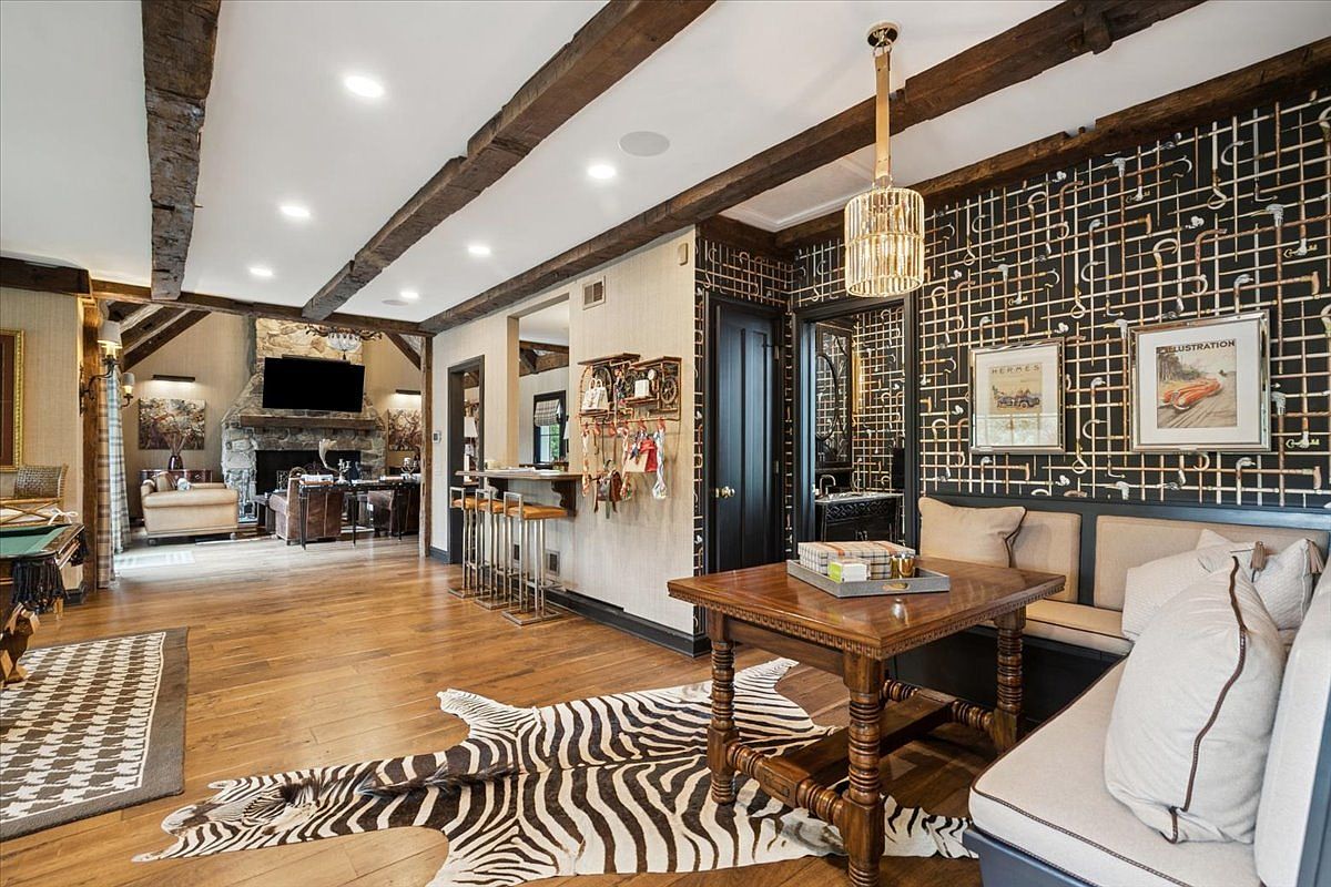 This expansive, open-concept living area features a rustic aesthetic with exposed dark wood ceiling beams and warm wood flooring. The space includes a cozy dining nook with a built-in bench, a bar area with high stools, and a view into a secondary living space anchored by a stone-clad chimney breast. A zebra-print rug adds a bold, eclectic touch to the room, which is illuminated by a decorative pendant light and recessed lighting.