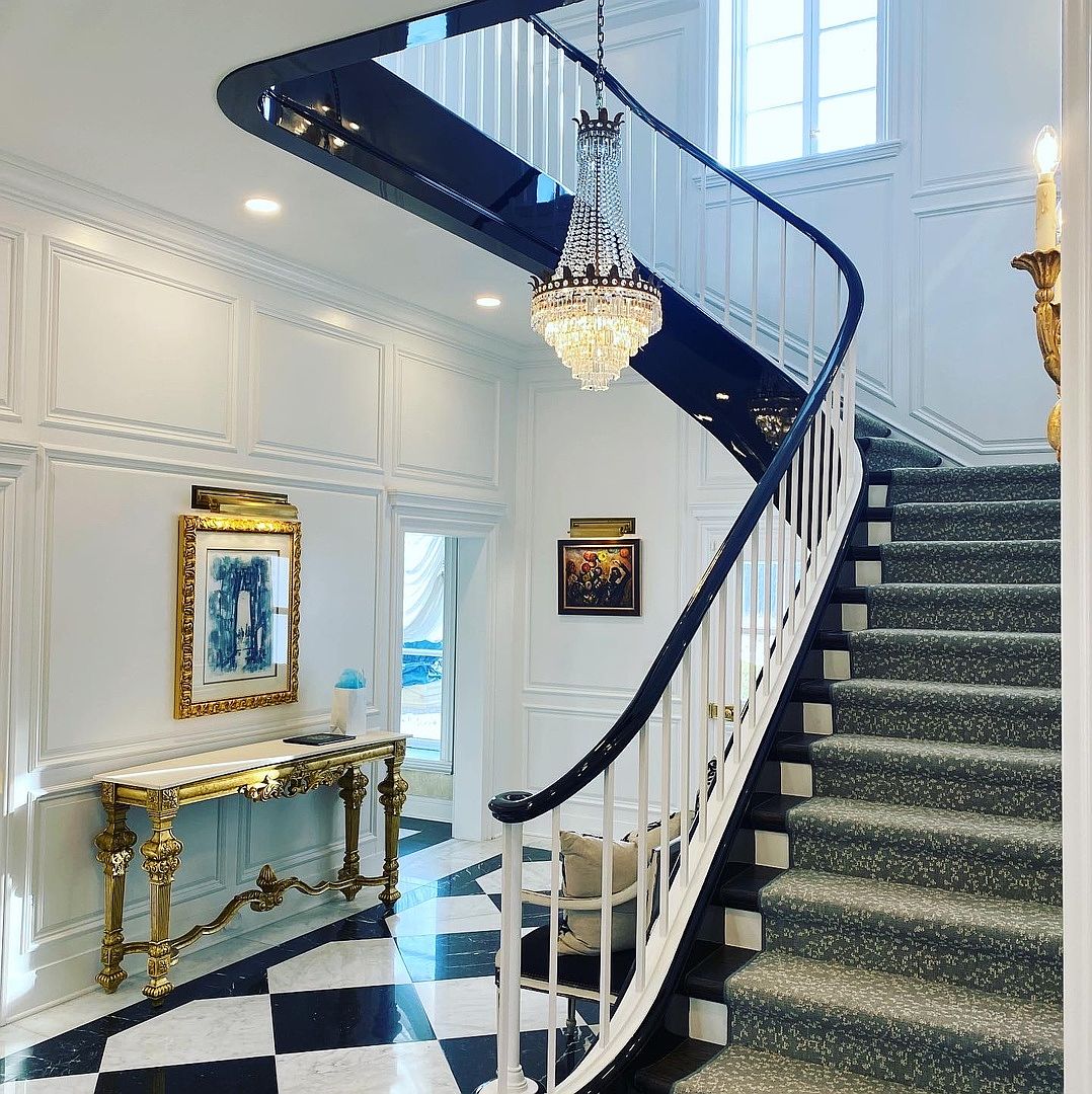This grand foyer features a dramatic curved staircase with a dark-finished handrail and white balusters, set against a backdrop of classic wainscoting. The floor is adorned with a bold black-and-white checkered pattern, complemented by a luxurious gold-toned console table and a sparkling crystal chandelier. The perspective captures the elegance of the entryway, highlighting the sophisticated architectural details and high-end finishes.