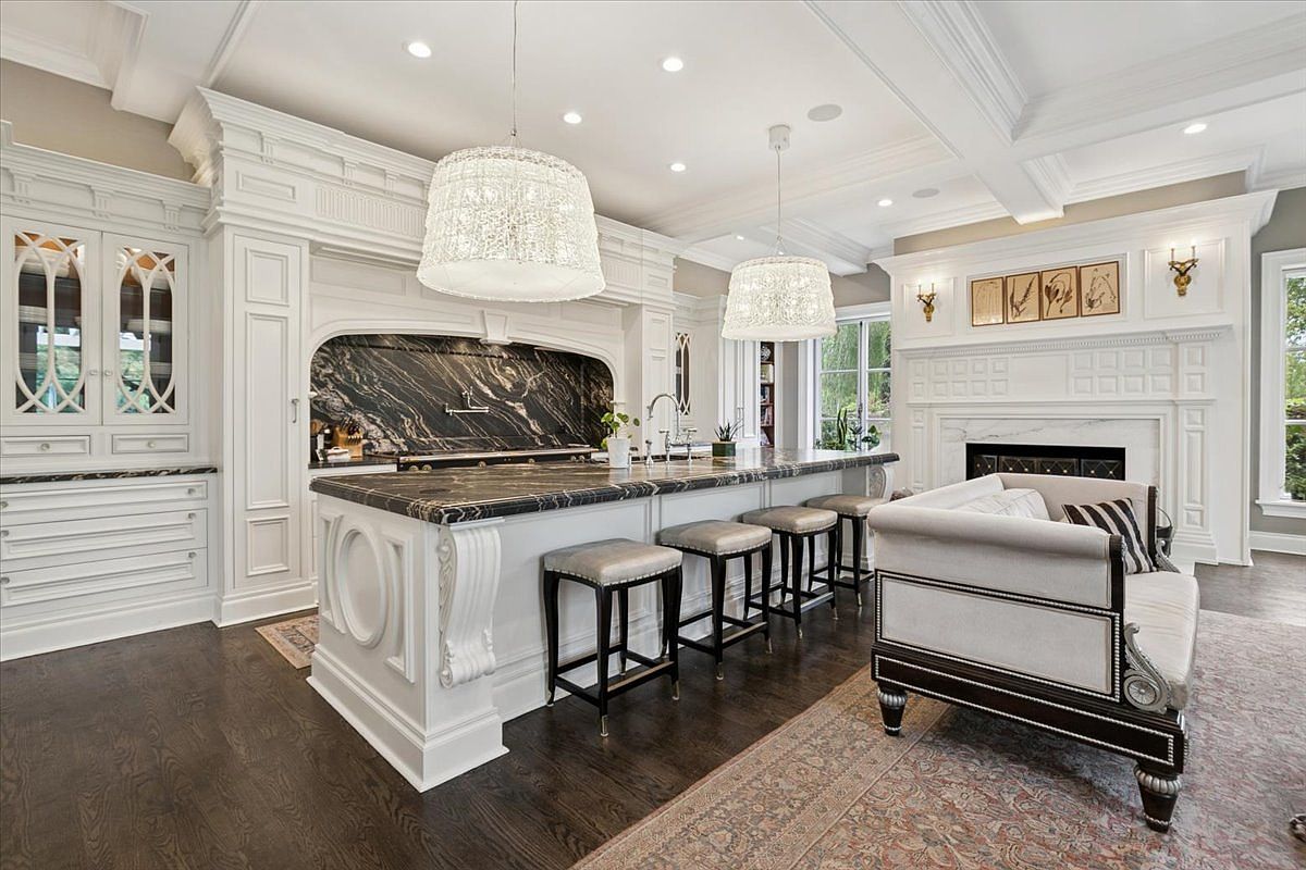 This elegant, open-concept kitchen features a large central island with a dark stone countertop and ornate white cabinetry. The space is illuminated by two large, textured pendant lights and includes a cozy seating area with a sofa positioned near a fireplace with a decorative white mantel. The room is finished with dark wood flooring and a neutral color palette, creating a sophisticated and inviting atmosphere.