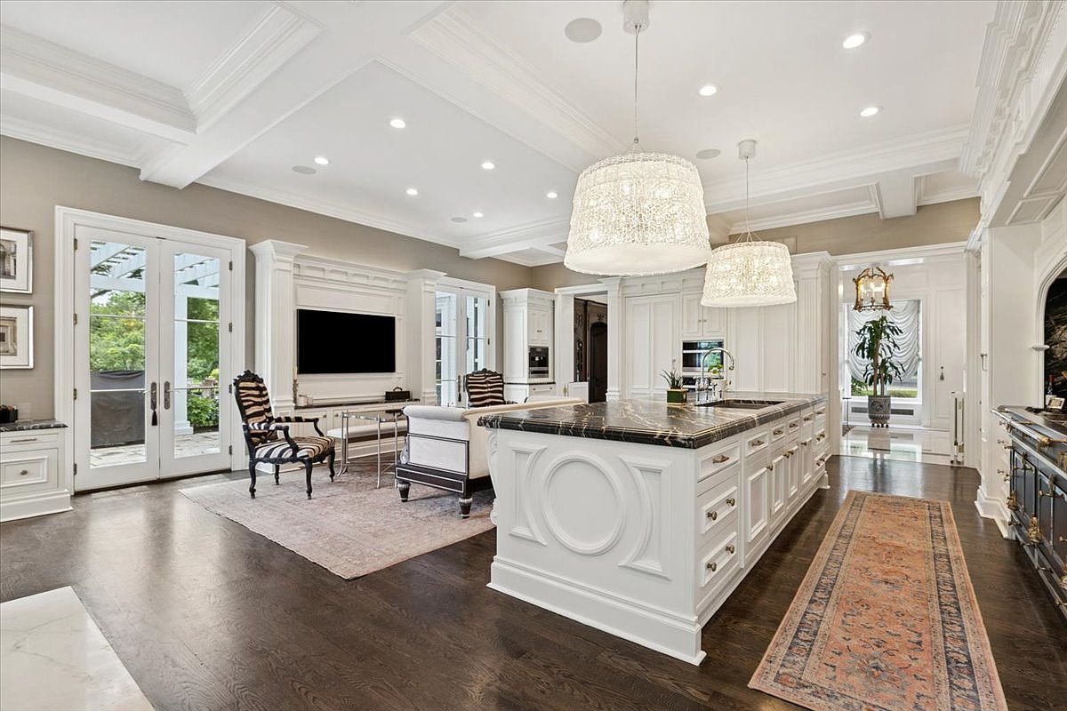 This expansive, open-concept kitchen features a large central island with a dark stone countertop and white cabinetry, complemented by dark hardwood flooring throughout. The space seamlessly integrates a living area with a television and patterned accent chairs, all illuminated by two large, textured pendant lights. The room is bright and airy, with tall glass doors providing a view of the outdoor greenery and a sophisticated, classic design aesthetic.