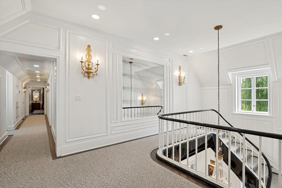 This elegant upper-level landing features crisp white wainscoting and walls, complemented by a patterned carpet that runs throughout the hallway. A grand staircase with a dark handrail and white balusters descends into the lower level, while ornate gold wall sconces and a large mirror add a touch of classic sophistication. The perspective captures the transition between the hallway and the stairwell, highlighting the bright, airy atmosphere of the home's interior architecture.