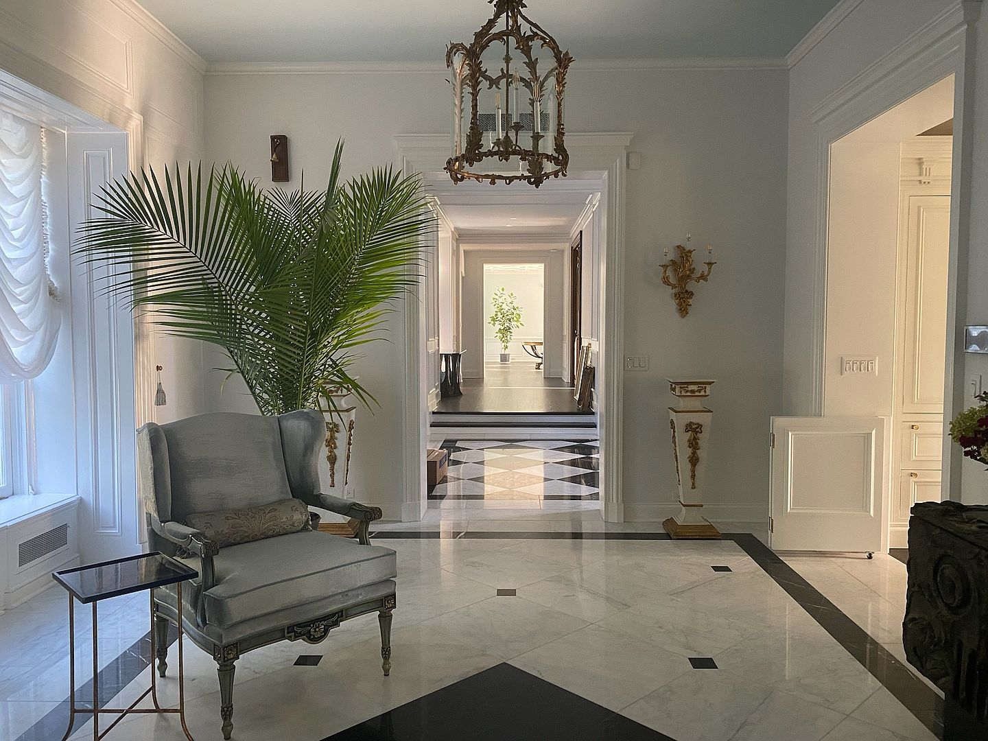 This elegant foyer features a sophisticated black and white patterned stone floor and a grand, ornate metal chandelier hanging from a light blue ceiling. A plush, light-colored wingback chair sits beside a large potted palm, while a long, deep hallway leads the eye toward a bright, sunlit room in the distance. The space is accented by classical wall sconces and decorative pedestals, creating a formal and luxurious atmosphere.
