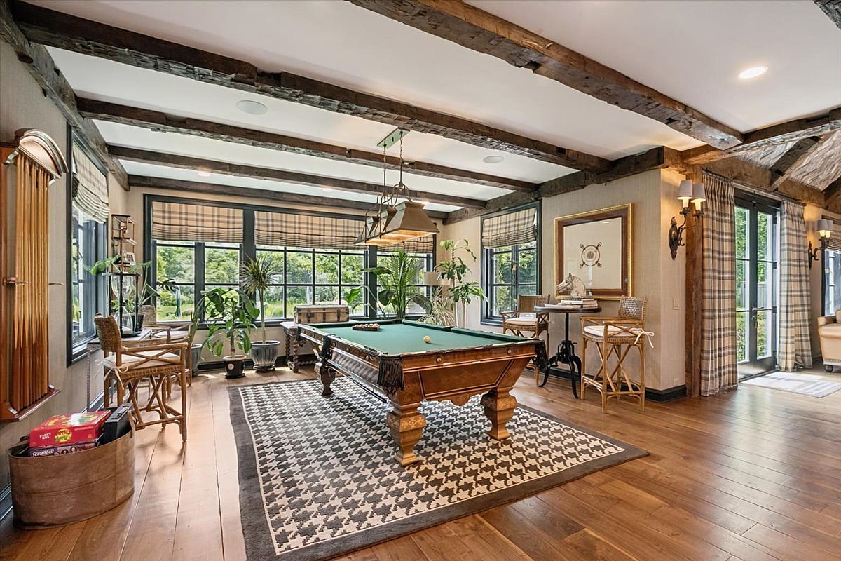 This elegant game room features a central pool table set atop a houndstooth-patterned area rug, framed by exposed dark wood ceiling beams. Large windows with plaid shades line the walls, bathing the space in natural light and offering views of the surrounding greenery. The room is accented with wicker-style chairs, a wall-mounted cue rack, and decorative wall art, creating a sophisticated yet relaxed atmosphere.