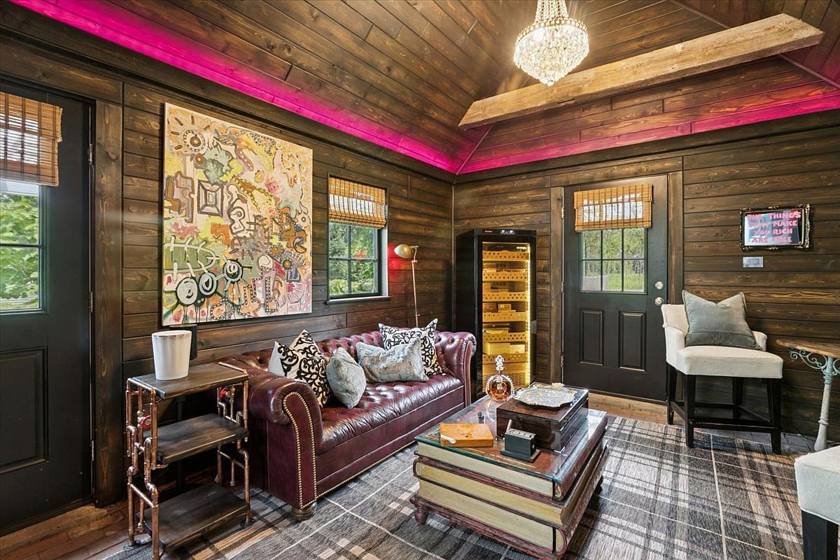 This cozy, rustic-inspired living space features dark wood-paneled walls accented by vibrant pink LED cove lighting. The room is furnished with a deep red leather sofa, a unique coffee table designed to look like a stack of vintage books, and a tall wine refrigerator. A large, colorful abstract painting serves as a focal point, while a crystal chandelier adds a touch of elegance to the warm, wood-toned interior.