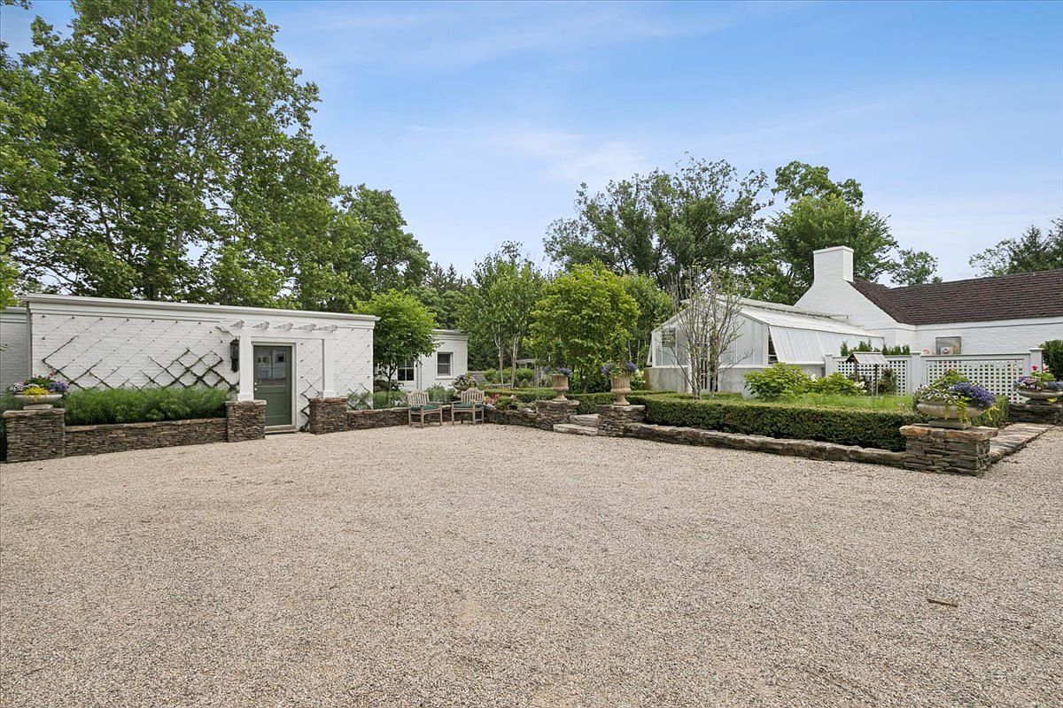 This serene outdoor space features a wide gravel courtyard framed by manicured hedges and stone-capped retaining walls. To the left, a white structure with a trellis-covered wall and a green door adds architectural interest, while a greenhouse with a white frame sits to the right. The scene is bathed in natural light, creating a peaceful and well-maintained garden atmosphere.