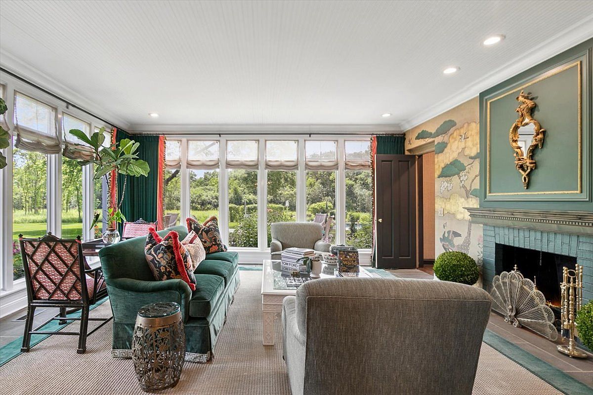 This elegant living room features a rich green velvet sofa and a pair of armchairs arranged around a central coffee table, creating a sophisticated seating area. A prominent fireplace with a decorative green tile surround and a gold-framed mirror serves as a focal point, complemented by a mural-style wall covering. Large windows line the back wall, flooding the space with natural light and offering a clear view of the lush greenery outside.
