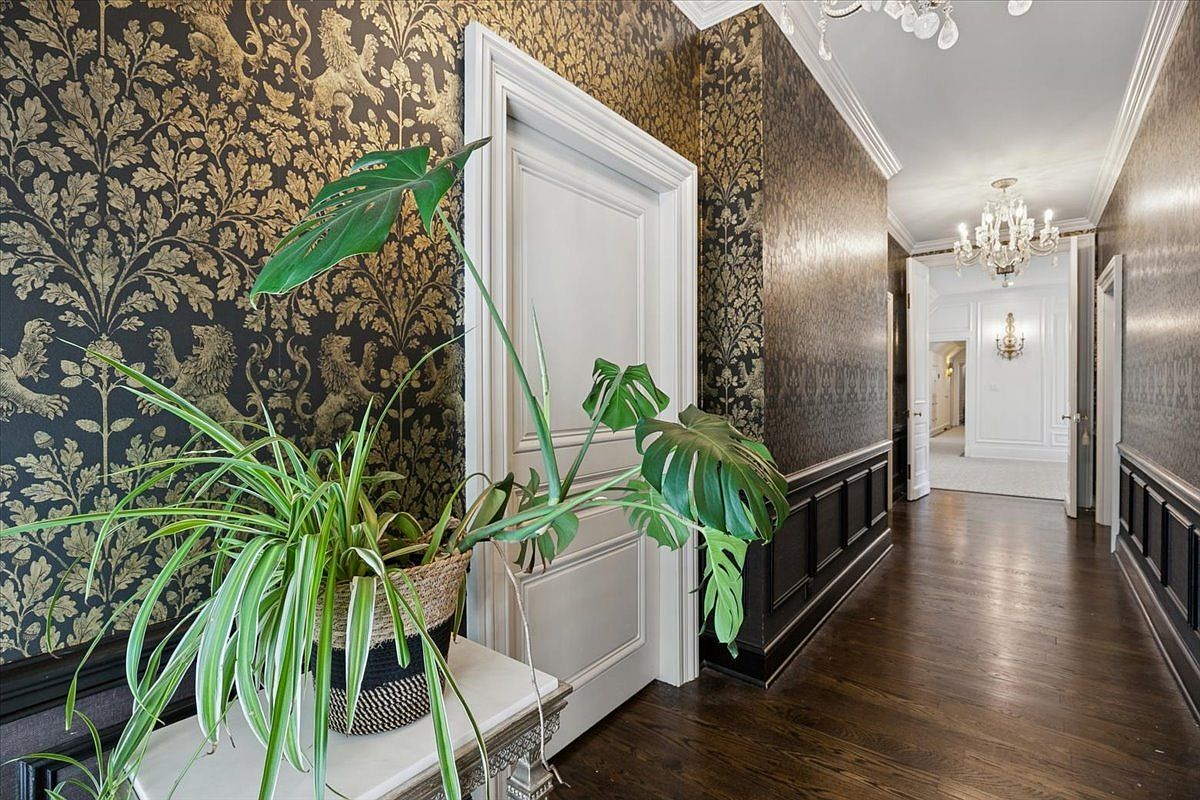 This elegant hallway features dramatic, dark-toned wallpaper with a gold botanical and lion motif, complemented by dark wood wainscoting and polished hardwood floors. A white door with classic molding stands out against the patterned walls, while a large potted plant on a console table adds a touch of greenery to the foreground. The perspective looks down the corridor toward an open doorway, revealing a bright, white-walled room illuminated by a crystal chandelier.