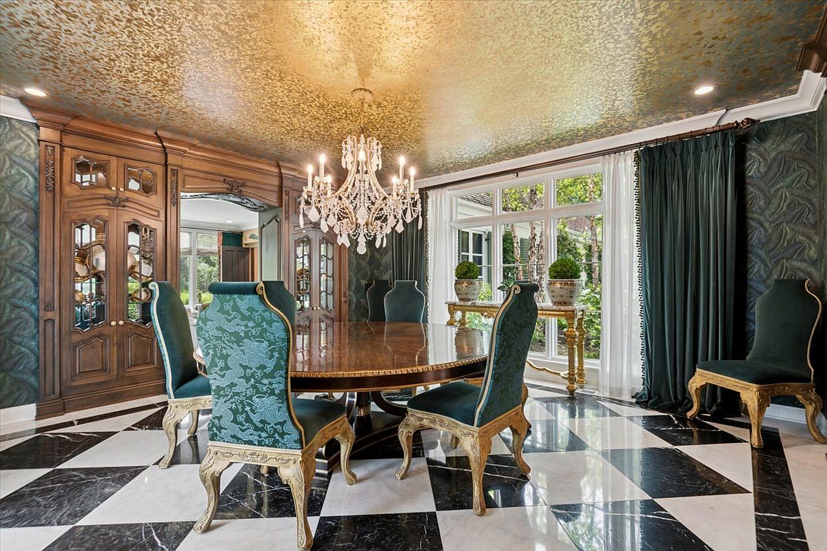 This opulent dining room features a large, round wooden table surrounded by ornate chairs with patterned upholstery and gilded frames. The space is defined by a striking black-and-white checkered stone floor, a dramatic gold-leaf ceiling, and deep green patterned wallpaper. A crystal chandelier serves as the centerpiece, while large windows draped in dark green curtains provide natural light and a view of the exterior greenery.