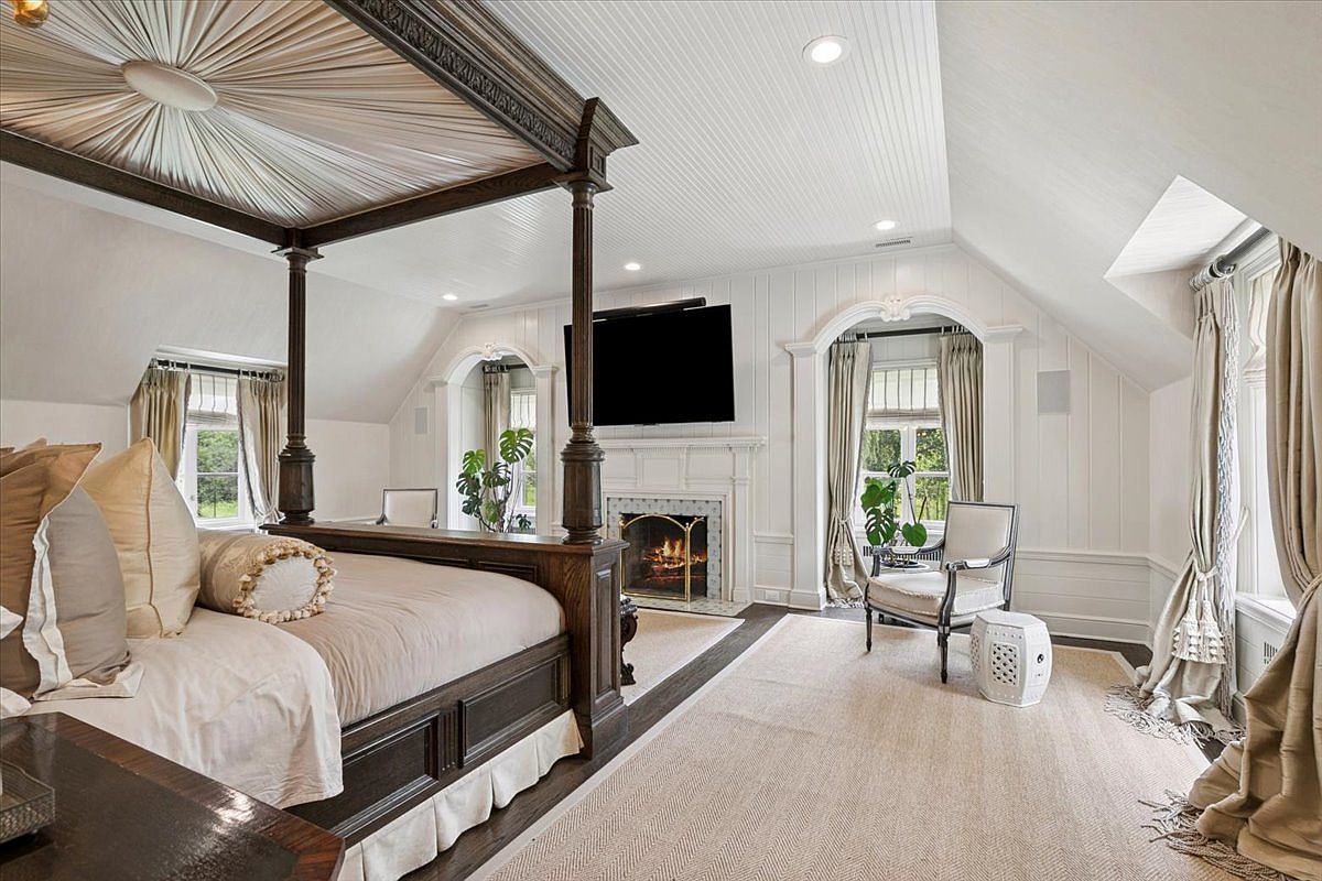 This elegant primary bedroom features a grand four-poster bed with a fabric-draped canopy as the central focal point. The room is characterized by white paneled walls, a vaulted ceiling, and a fireplace with a decorative mantel and television mounted above. Large windows with soft drapery flank the fireplace, allowing natural light to fill the space, while a comfortable armchair and a patterned rug complete the sophisticated, tranquil atmosphere.