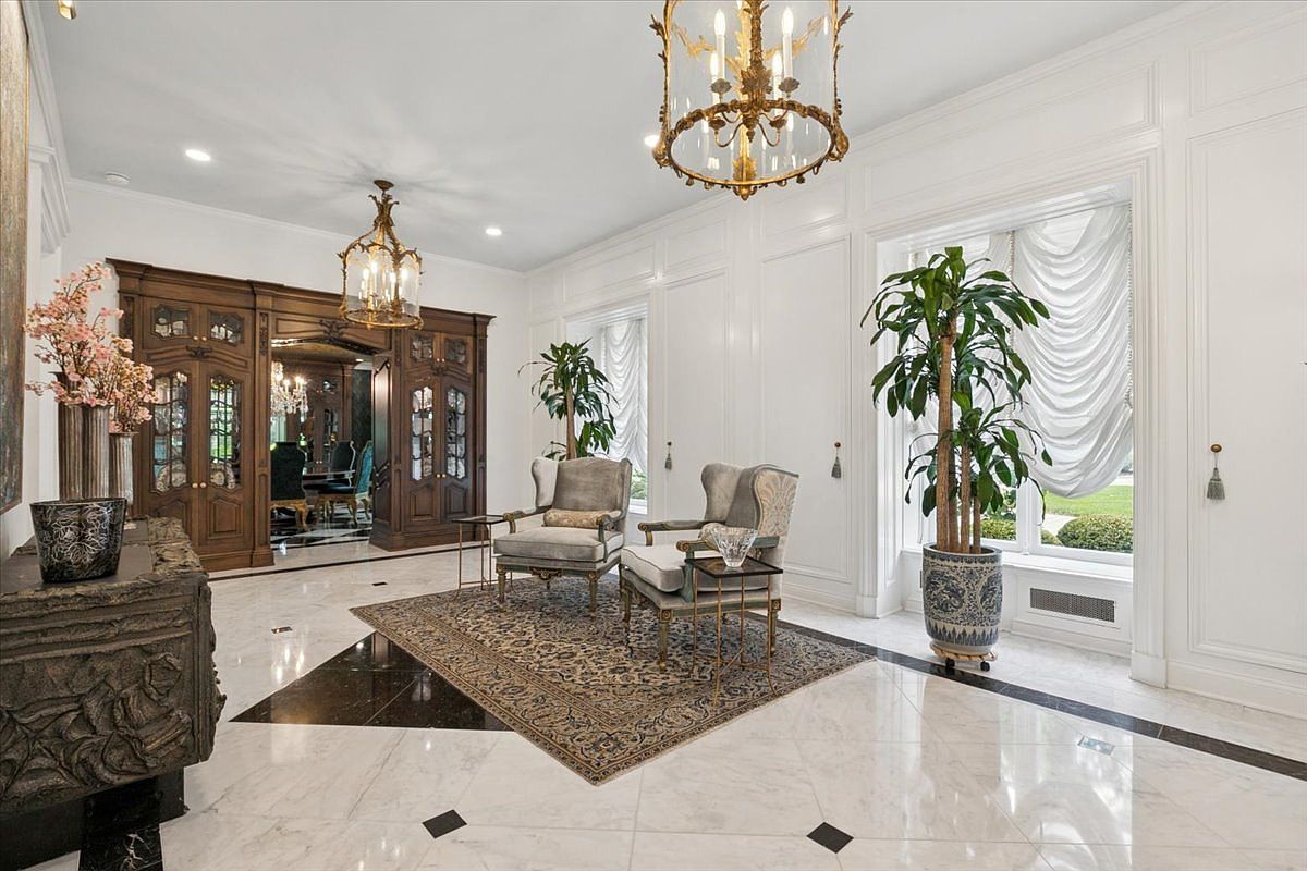 This elegant foyer features polished stone flooring with dark geometric accents and a central area rug beneath two classic armchairs. The space is illuminated by ornate gold chandeliers and features tall windows draped in soft, white fabric, while a dark wood built-in cabinet adds a rich contrast to the bright, white-paneled walls. The perspective is a wide, eye-level shot that captures the sophisticated, formal atmosphere of the home's entrance.