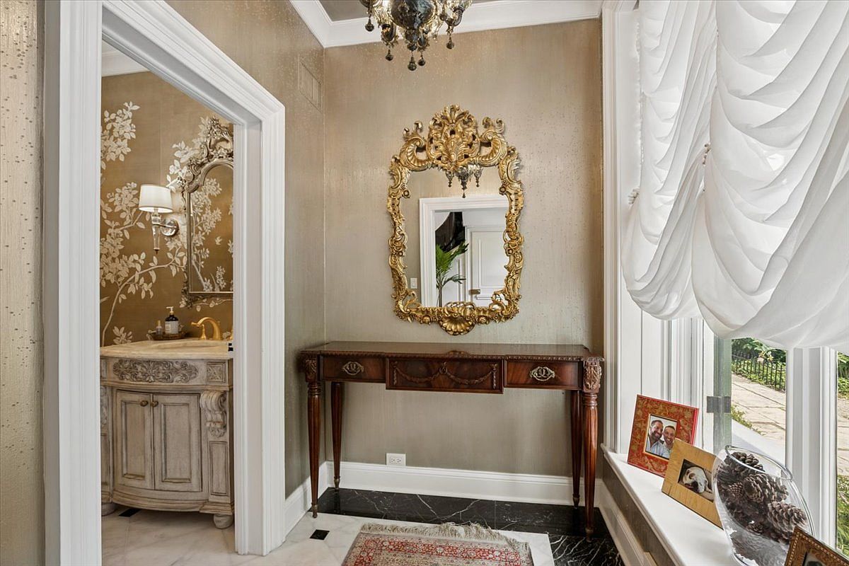 This elegant entryway features a console table topped with an ornate gold-framed mirror, set against a shimmering metallic wall finish. To the left, a doorway leads into a powder room decorated with floral wallpaper and a stone-topped vanity, while to the right, large windows draped in soft white curtains allow natural light to fill the space. The floor transitions from a patterned tile in the hallway to a dark stone border, creating a sophisticated and welcoming atmosphere.