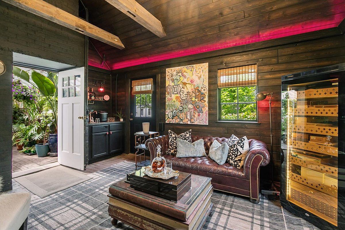 This cozy, rustic-inspired lounge features dark wood-paneled walls, a tufted leather sofa, and a plaid-patterned rug, creating a sophisticated, club-like atmosphere. A large, abstract painting serves as a focal point above the seating area, while a glass-fronted humidor and a small wet bar area add to the room's refined character. The space opens directly into a lush, sunlit conservatory, providing a seamless transition between the moody interior and the vibrant greenery outside.