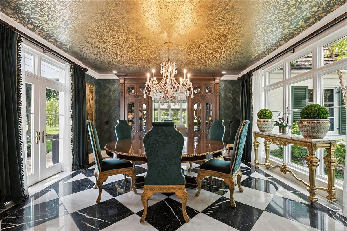 This opulent dining room features a large circular table surrounded by high-backed chairs upholstered in deep green fabric with ornate gold-toned legs. The space is defined by a striking black and white checkered stone floor, a metallic gold-leaf ceiling, and a grand crystal chandelier that serves as the focal point. Large windows and glass doors allow for abundant natural light, while a decorative console table with potted greenery adds a touch of elegance to the perimeter.