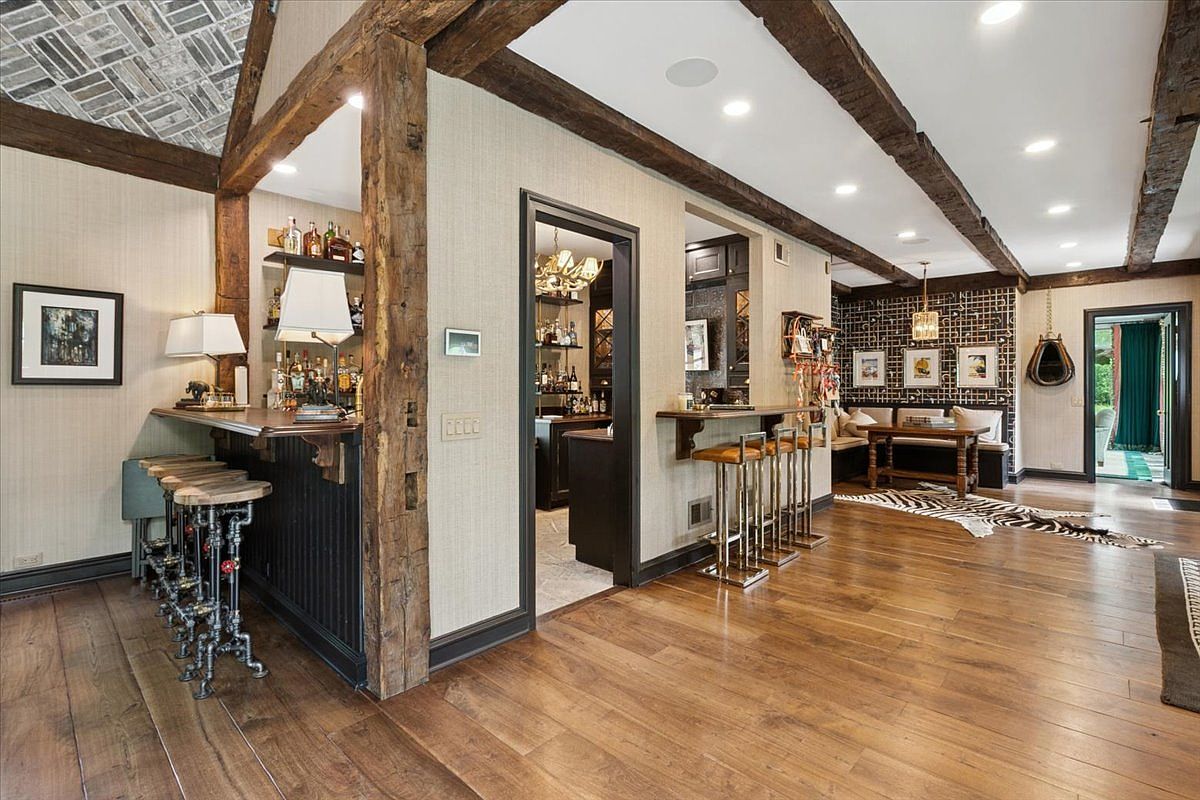 This expansive basement area features a rustic, lodge-inspired aesthetic with exposed timber beams and warm wood flooring. The space includes a dedicated bar area with industrial-style stools, a secondary walk-through bar, and a cozy seating nook with a bench and table. The room is illuminated by recessed lighting and decorative fixtures, creating a sophisticated yet comfortable atmosphere for entertaining.