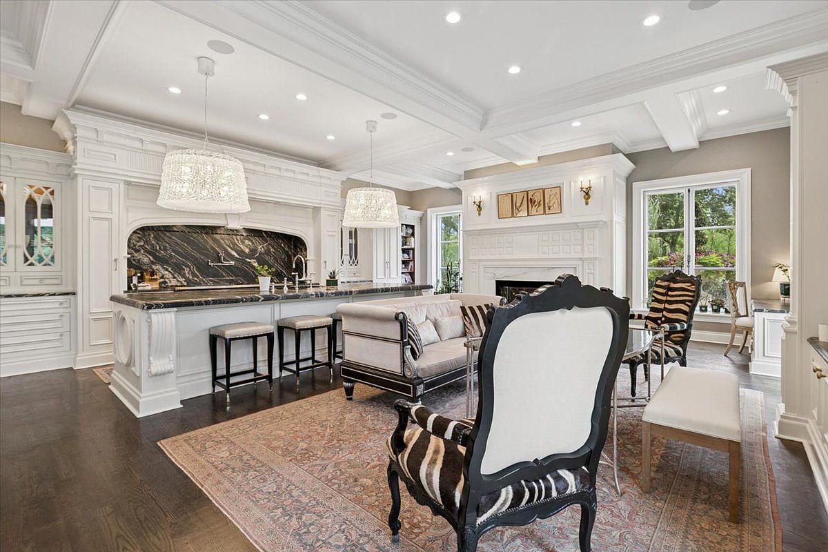 This expansive open-concept kitchen and living area features a large central island with a dark, veined stone countertop and bar seating. The space is anchored by a sophisticated fireplace with a white mantel and a decorative surround, complemented by elegant furniture including zebra-print accent chairs. The room is illuminated by dual crystal pendant lights and recessed lighting, creating a luxurious and cohesive living environment.