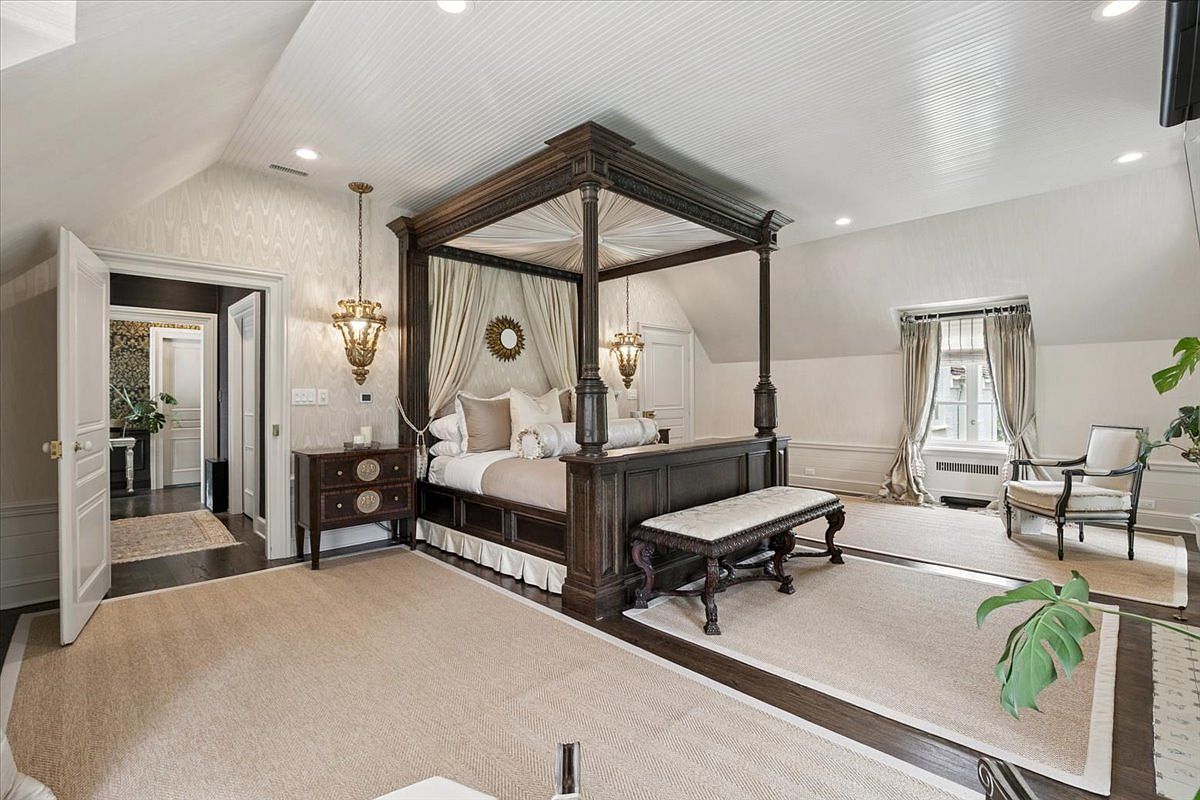 This elegant primary bedroom features a grand four-poster canopy bed as the central focal point, draped in soft fabric and positioned on a large area rug. The room is characterized by its sophisticated, traditional aesthetic, including patterned wallpaper, ornate hanging light fixtures, and a classic bench at the foot of the bed. A cozy seating area with a chair is situated near the window, while a doorway leads to an adjacent room, creating a sense of spaciousness and luxury.