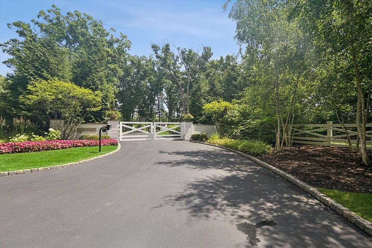 This image captures the gated entrance to a private estate, featuring a paved driveway leading up to a white wooden gate flanked by stone pillars. The property is surrounded by lush, mature trees and vibrant pink flower beds, creating a secluded and welcoming atmosphere. The perspective is a ground-level view looking directly toward the entrance, emphasizing the sense of privacy and prestige.