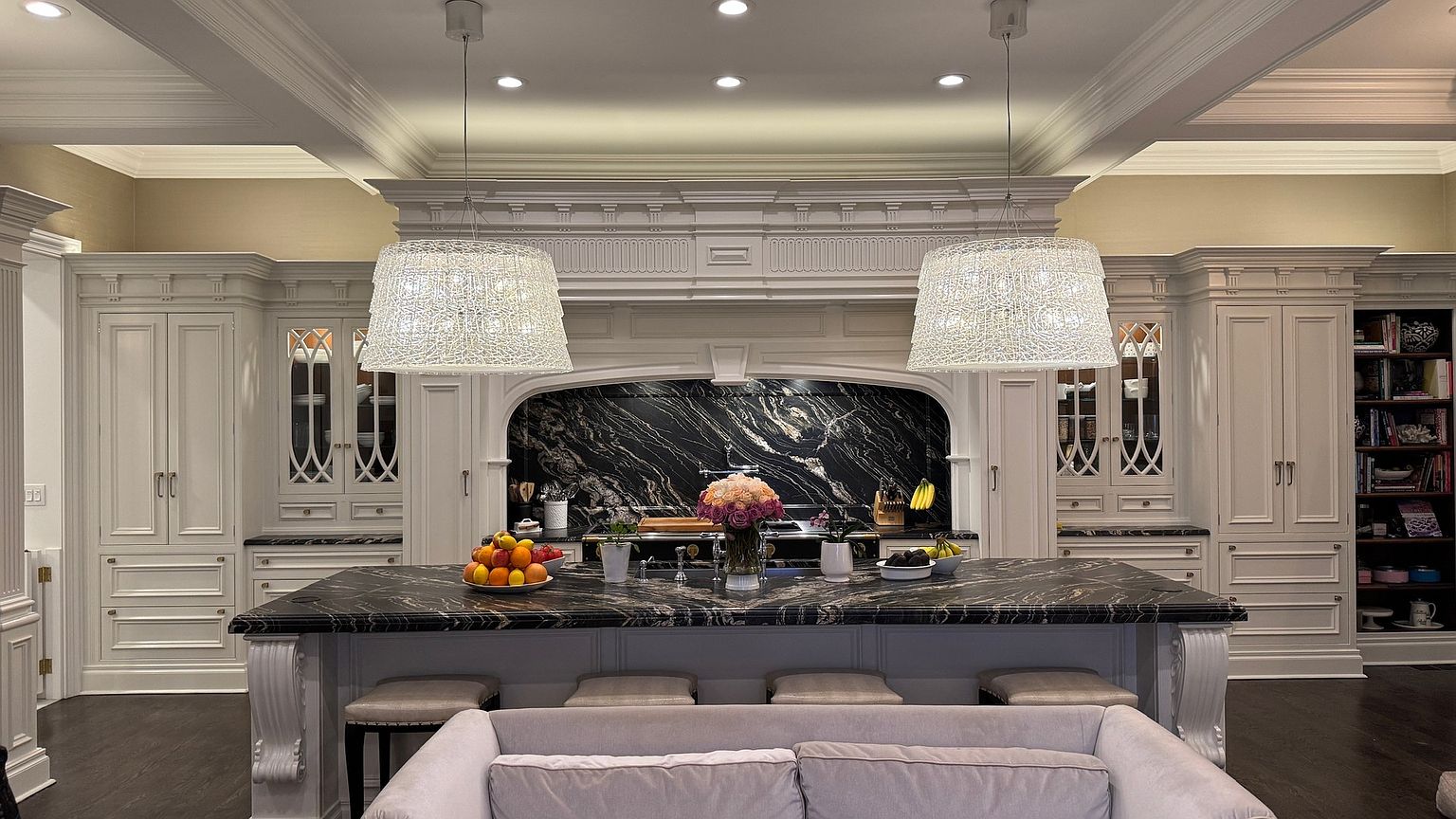 This sophisticated kitchen features a large central island with a dark, veined stone countertop and integrated seating, serving as the focal point of the room. The space is framed by elegant, light-toned cabinetry, a dramatic dark stone backsplash, and two large, textured pendant lights hanging above the island. The perspective is centered and symmetrical, highlighting the high-end finishes and the seamless integration of a cozy seating area in the foreground.